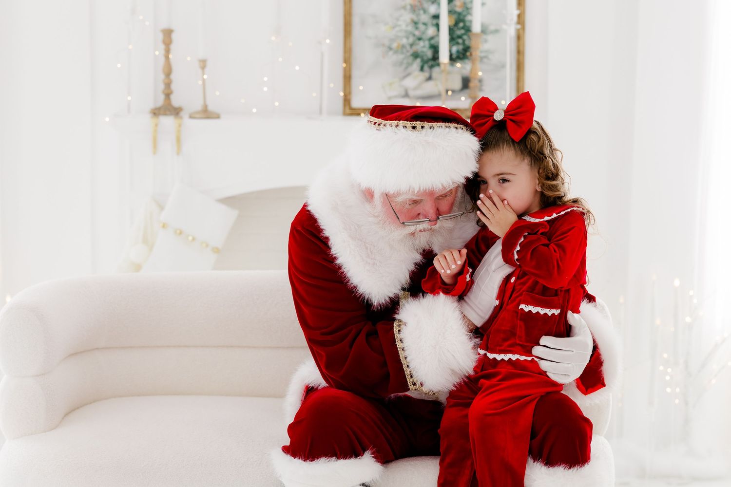 A little girl in red outfit whispers to Santa Claus on a white couch in a bright holiday photo setting.