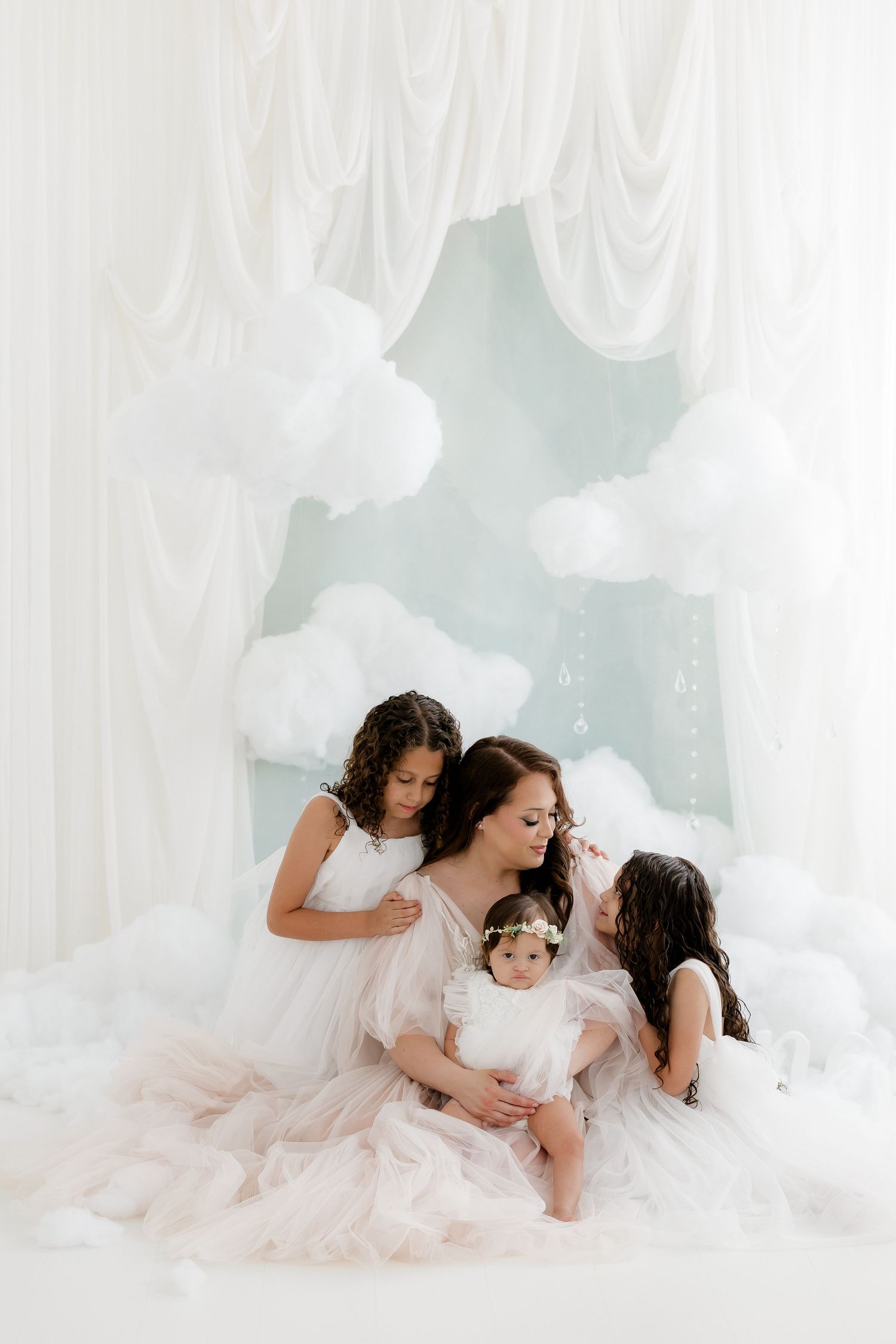 A mother tenderly kisses her baby while two daughters in white tulle dresses gather close in a cloud-themed studio.