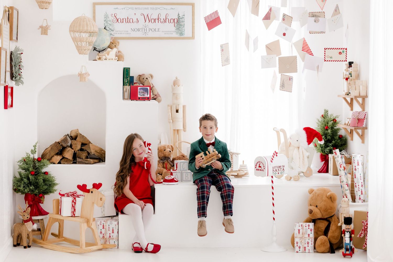 Two children sit in a festive Santa's Workshop Christmas backdrop with gifts, wooden toys, and holiday decorations.