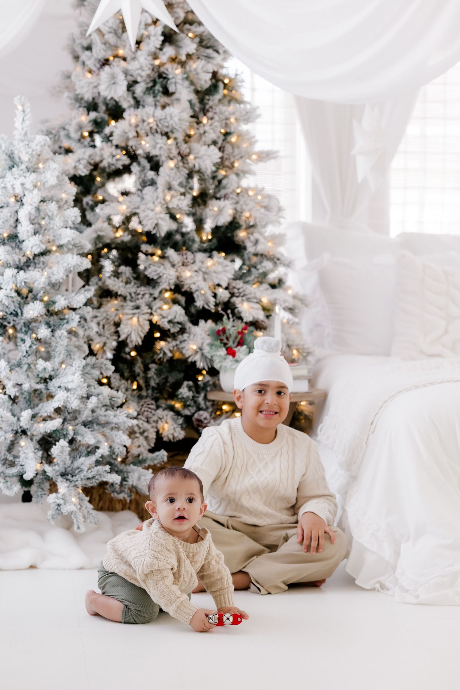 Two children in cream sweaters sit before a snow-flocked Christmas tree with warm lights in a white decorated room.