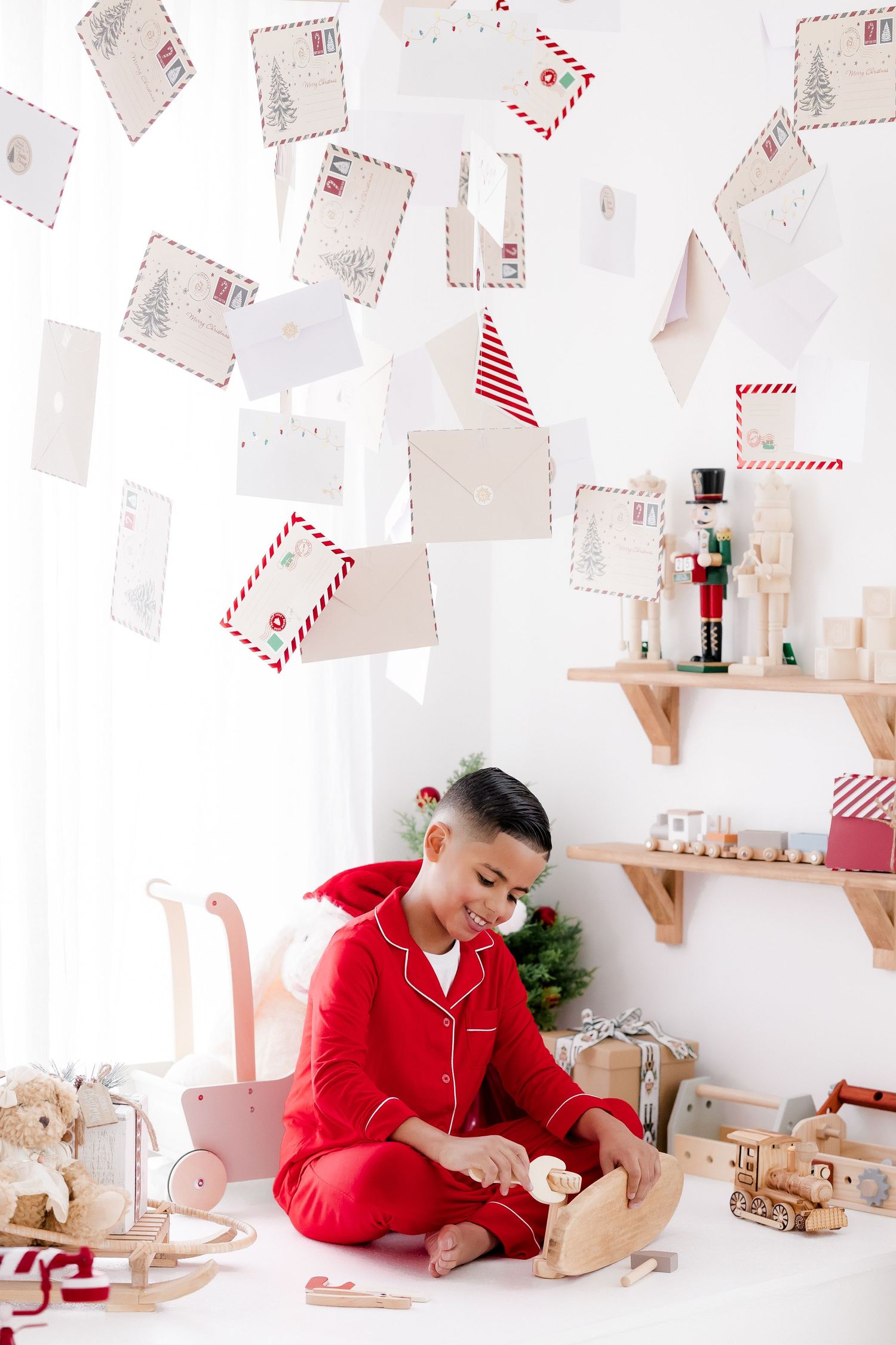 Child in red pajamas playing with wooden toys surrounded by Christmas decorations and hanging festive envelopes.