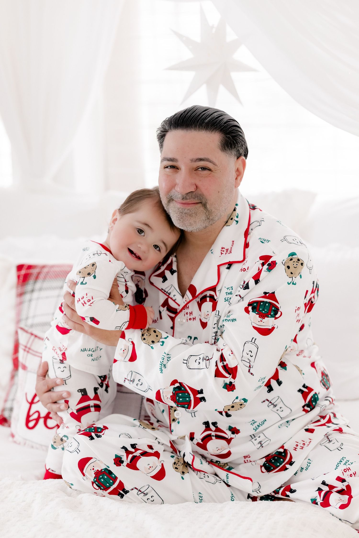 Father and child wearing matching Santa Claus Christmas pajamas sitting together on a white bed.