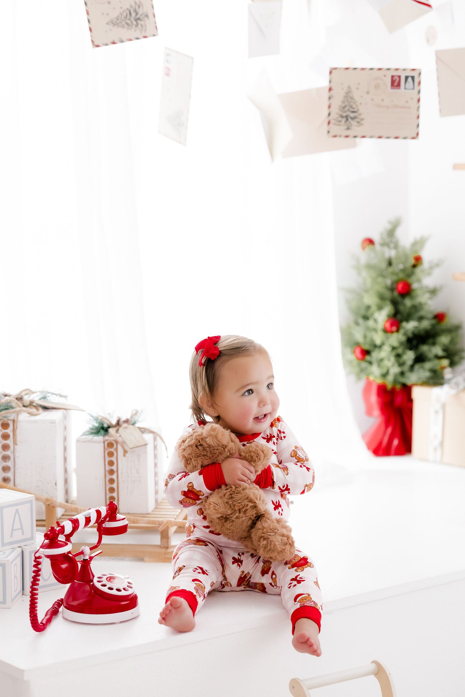 Toddler girl in Christmas pajamas hugging teddy bear surrounded by gifts and holiday decorations.