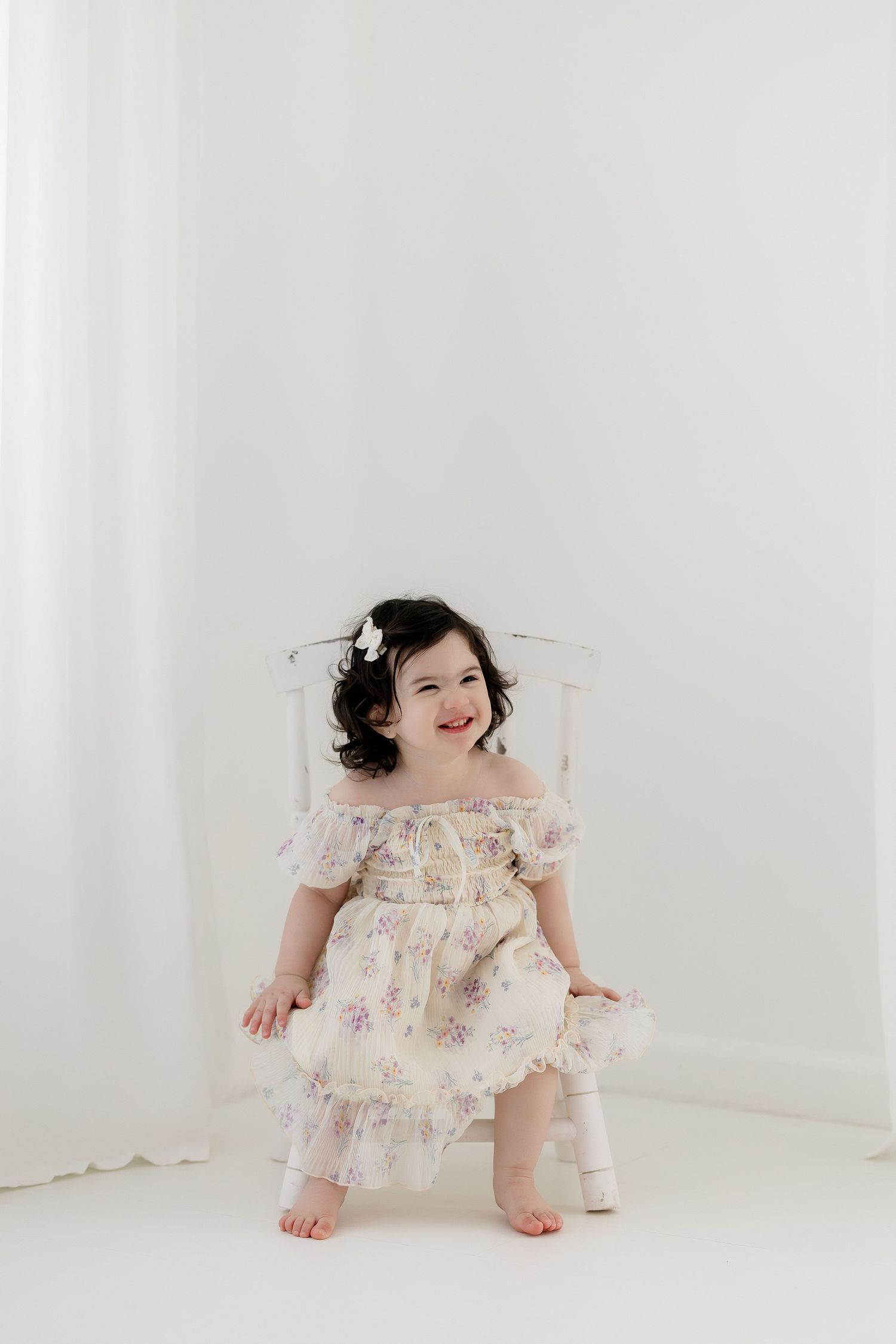 A smiling toddler girl in a floral ruffle dress with a hair clip sits on a white chair in a bright studio.