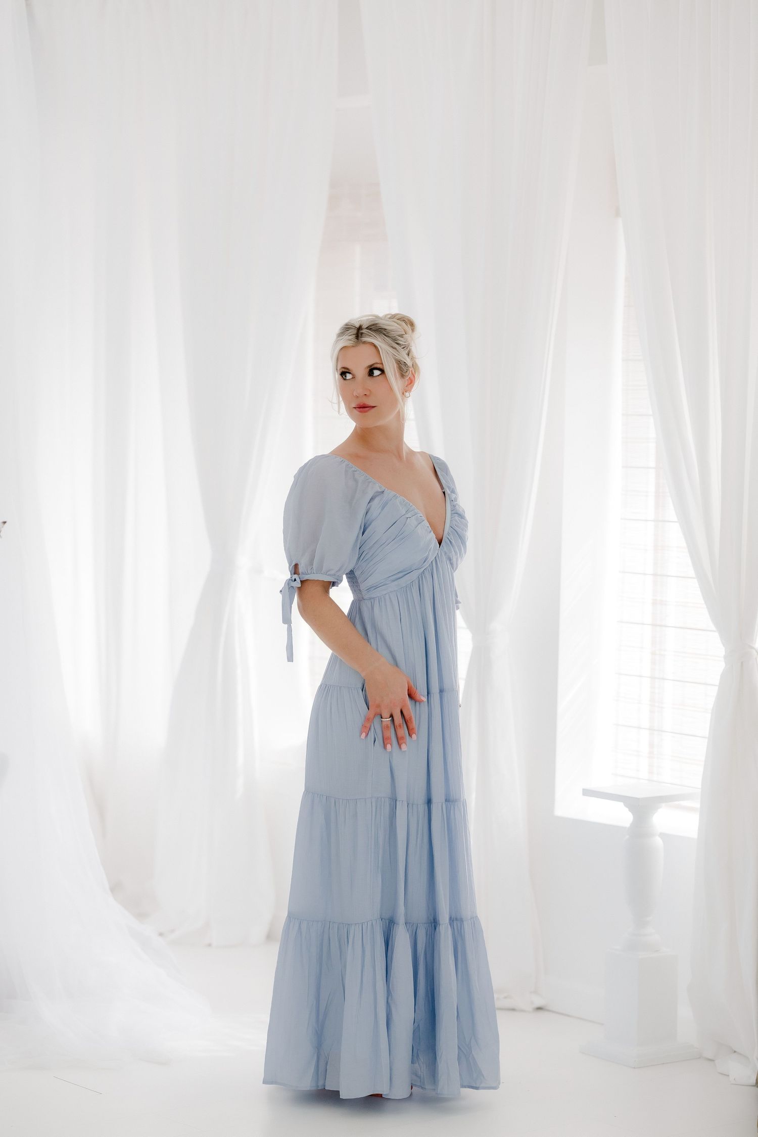 A person wearing a light blue tiered maxi dress poses in a bright white studio setting with flowing curtains.