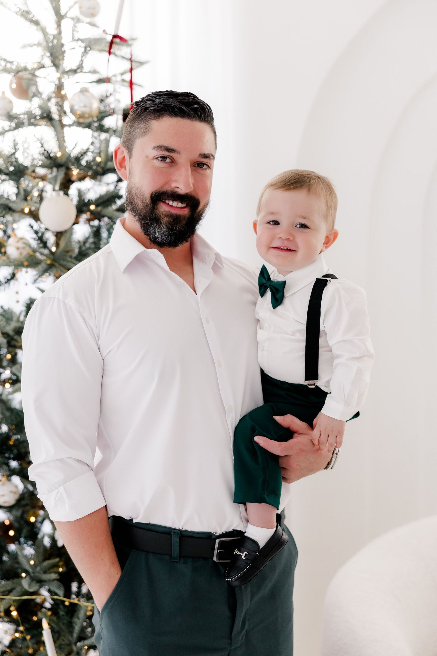 Father and child in formal attire share playful moments against white Christmas backdrop.