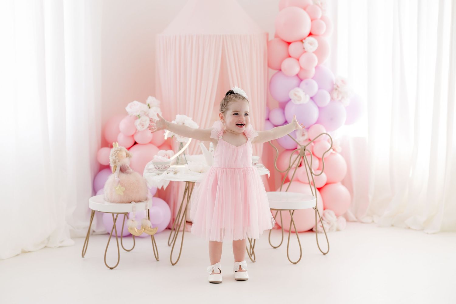 A pink and white themed birthday party setup with balloons, tulle decorations, and a small chair for a cake smash session.