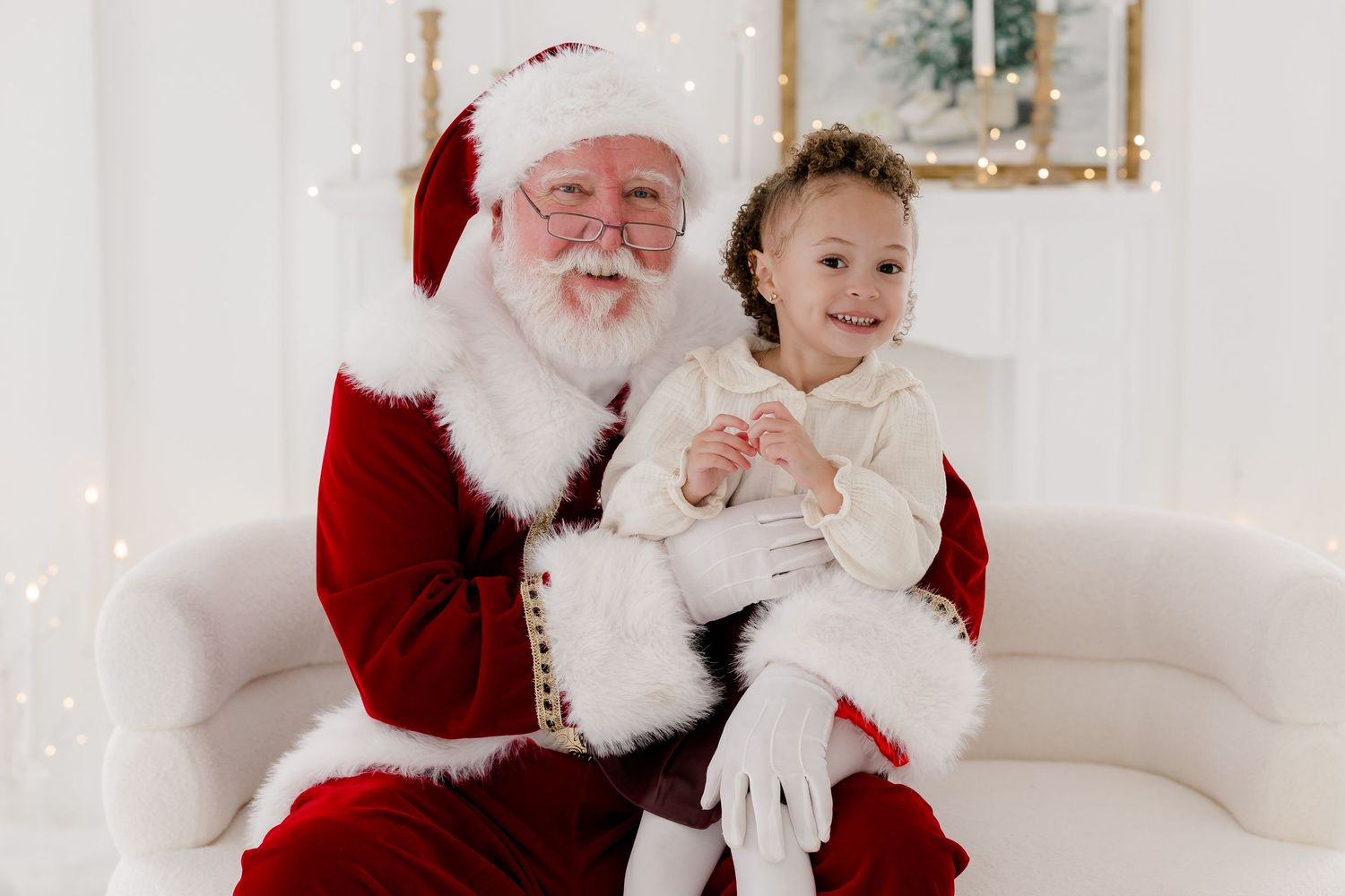 A young girl in a white dress sits on Santa Claus's lap, smiling together in a festive white holiday photo setting.