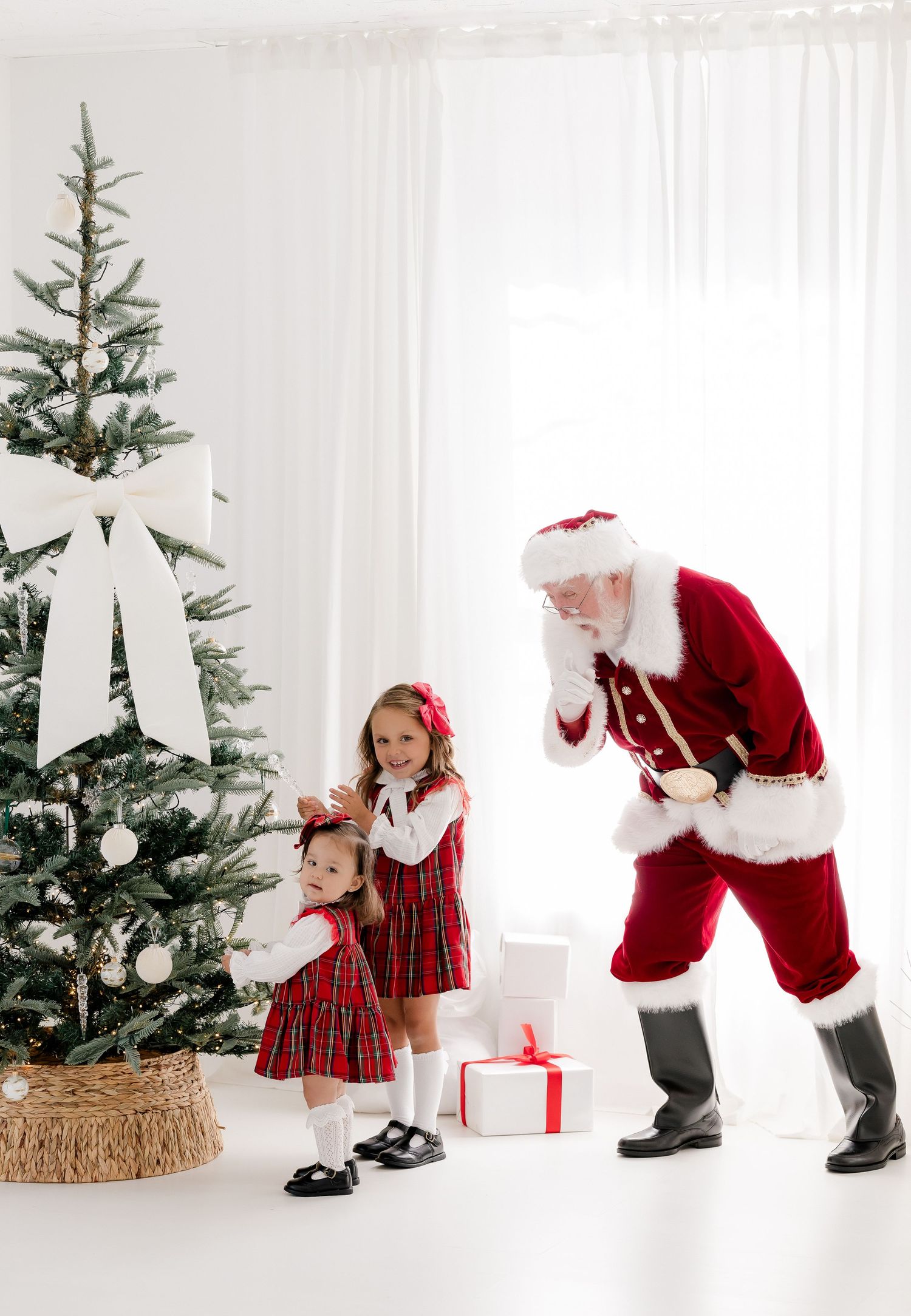 Two young girls in red plaid dresses receive gifts from Santa Claus beside a decorated Christmas tree.