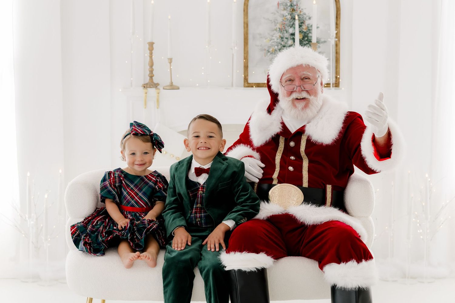 Two children in festive attire sit smiling beside Santa Claus on a white chair in a bright holiday photo studio.