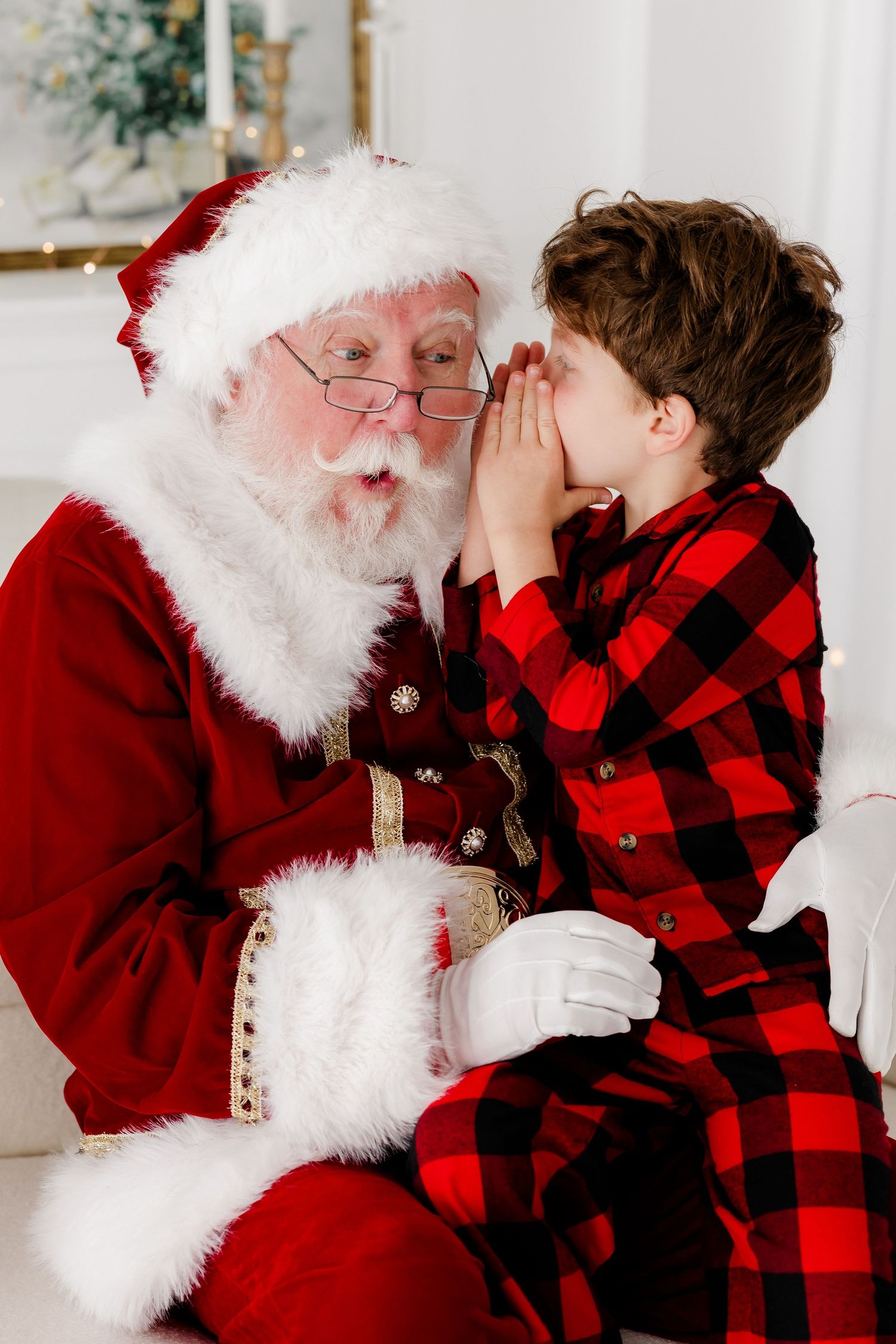 Young boy in plaid pajamas whispering his Christmas wishes to Santa Claus sitting together on a white couch.