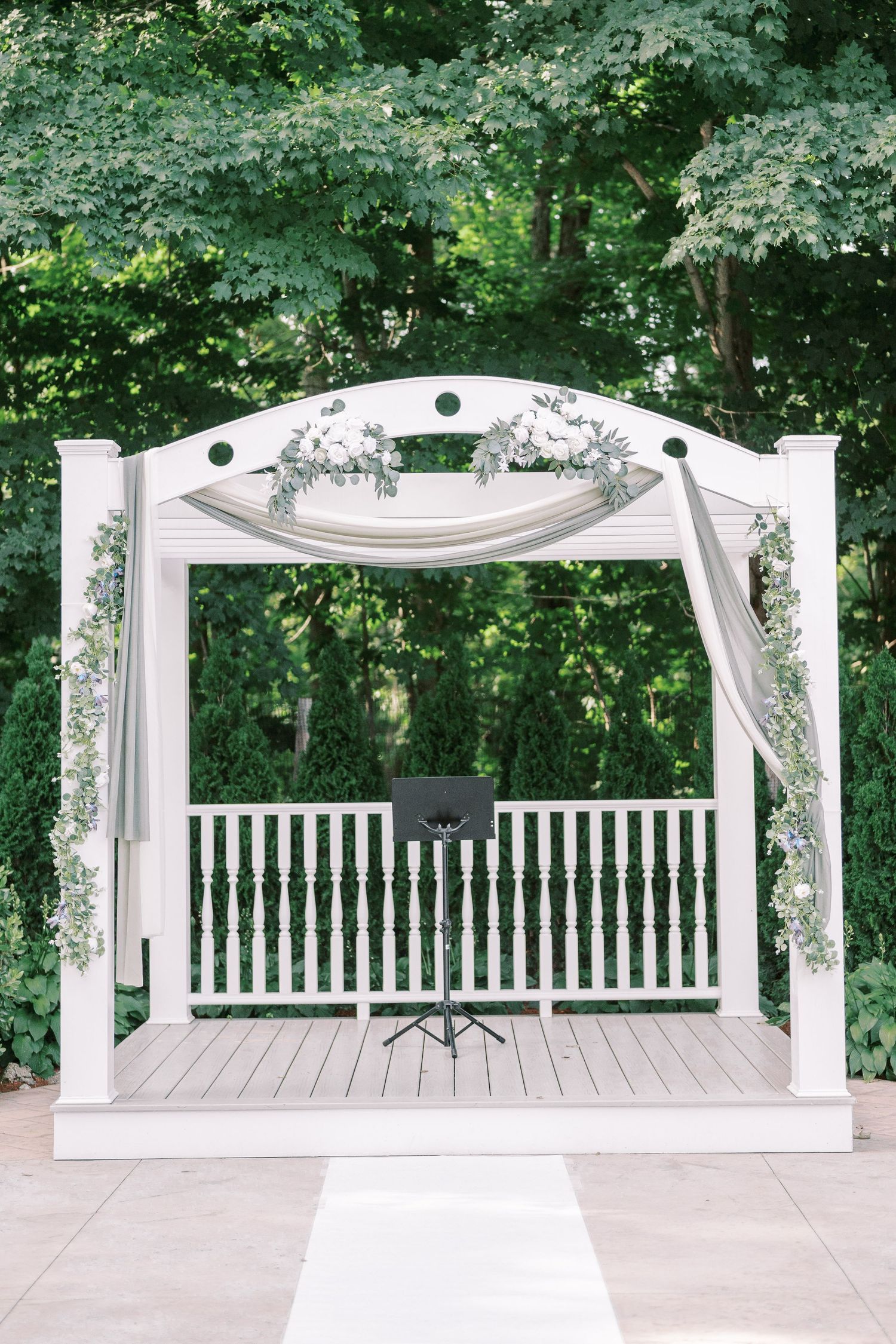 A white wooden wedding arch with floral decorations and draped fabric stands in front of a lush forest backdrop.