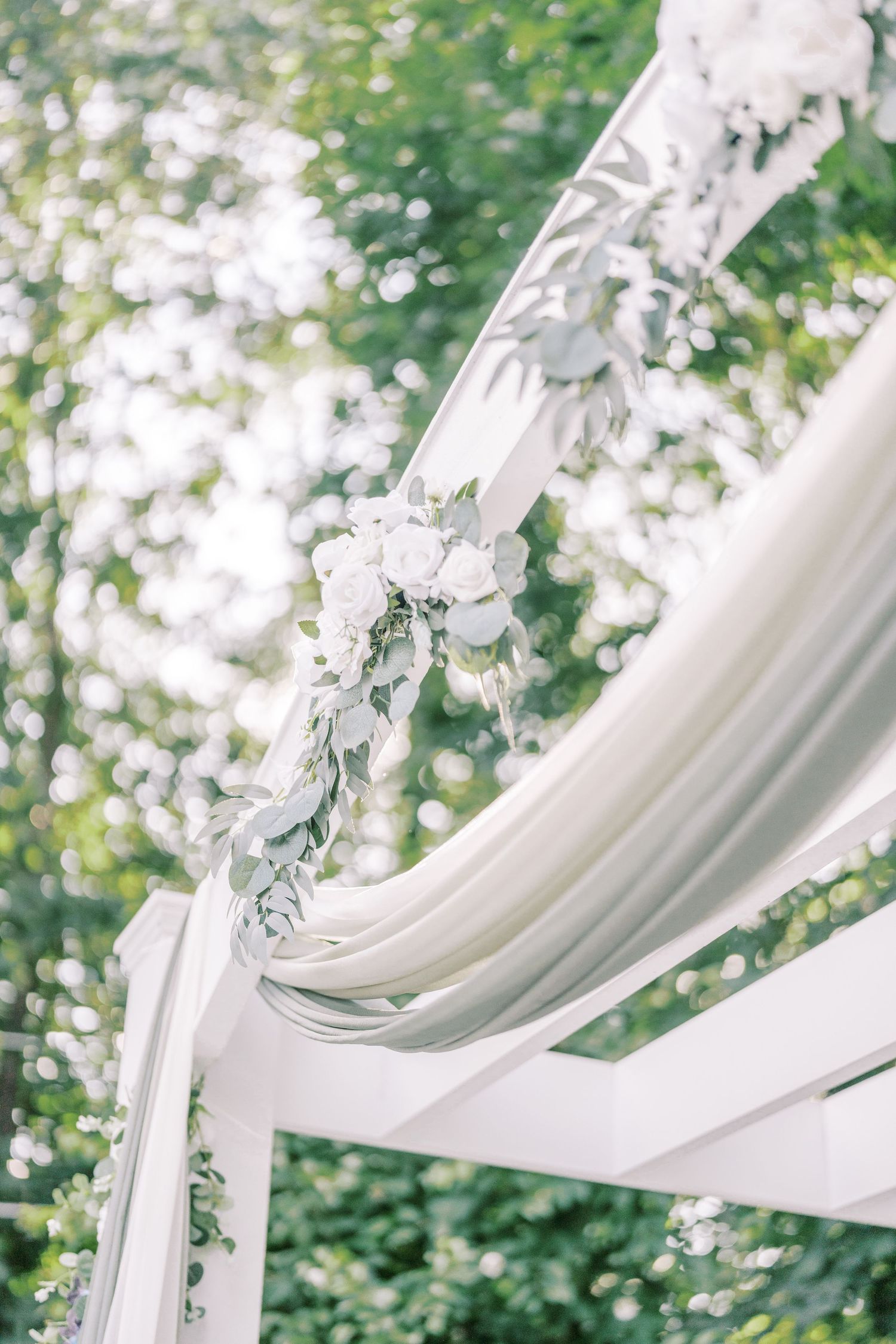White fabric drapes and floral decorations adorn an elegant outdoor wedding arch against a natural green backdrop.