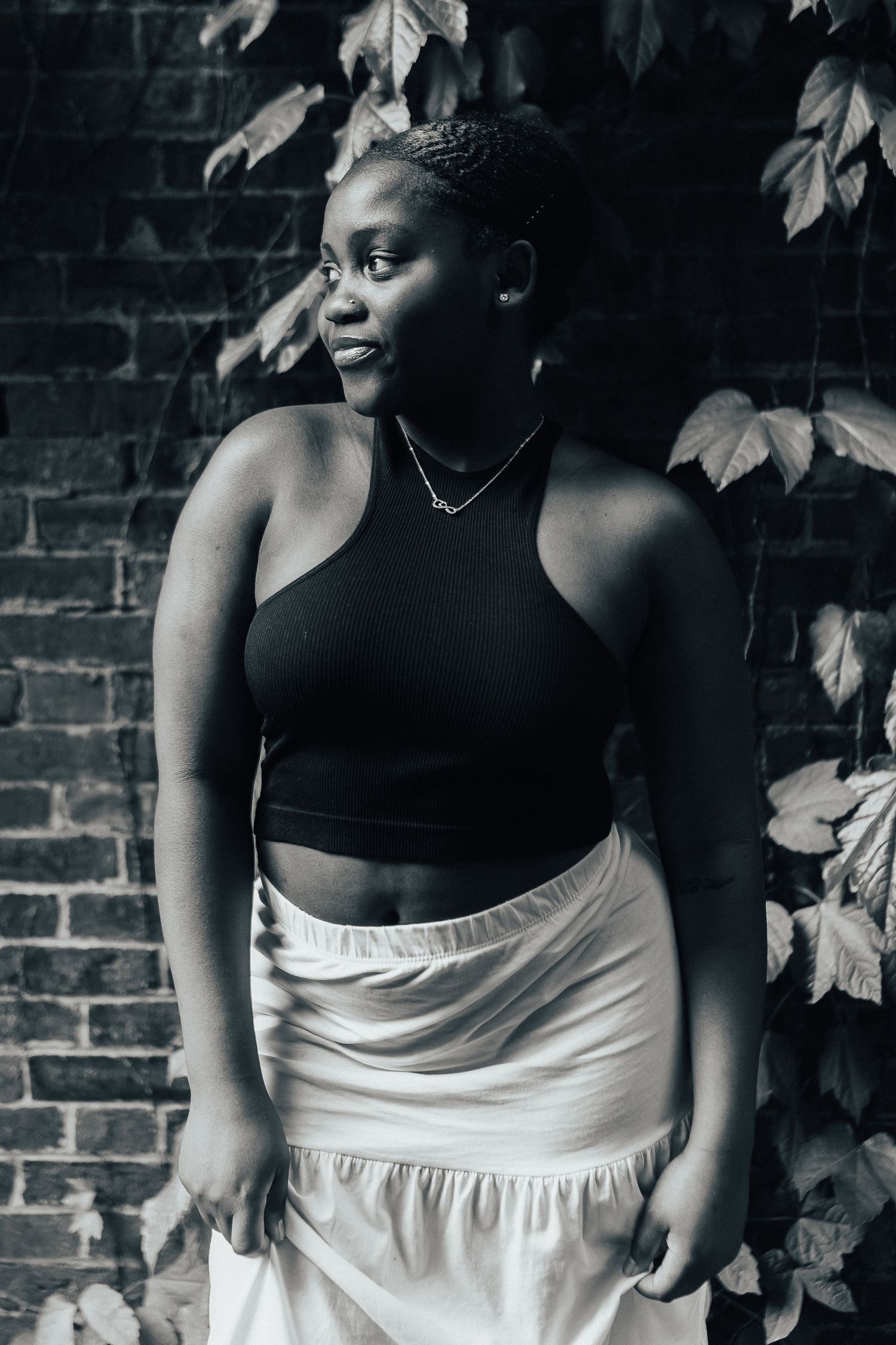 Black and white artistic portrait featuring a model in a black crop top and white skirt against a brick wall with leaves.