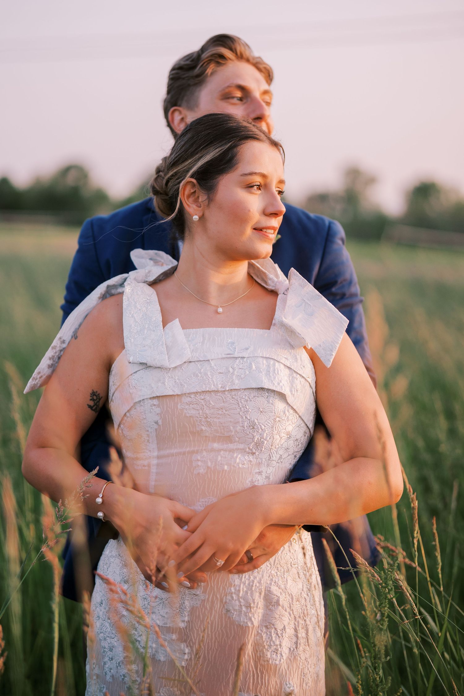 A couple embraces in a golden field at sunset, with the person in white wearing a detailed lace dress.
