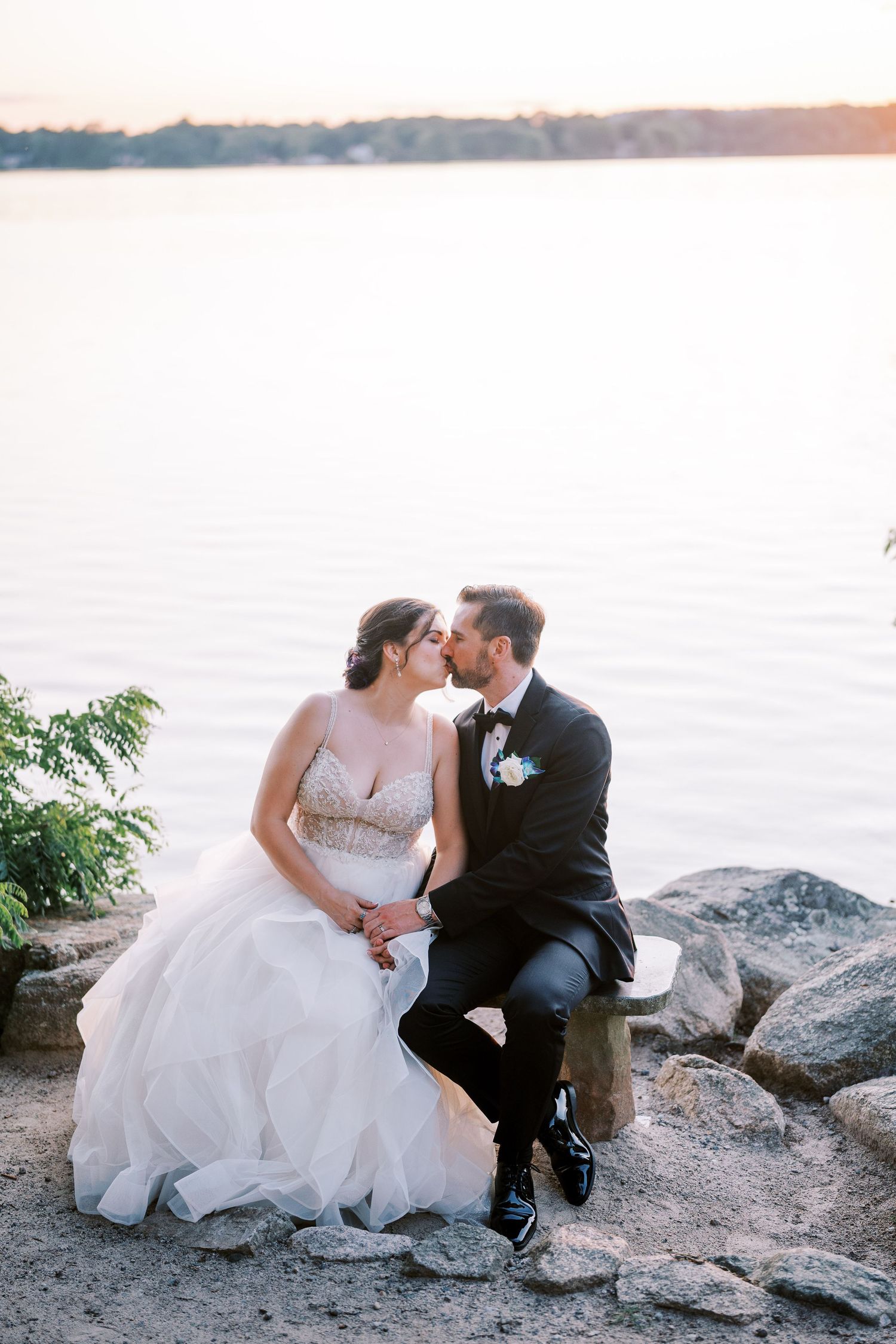 A couple sits together on coastal rocks at sunset while wearing formal wedding attire against a serene waterfront backdrop.