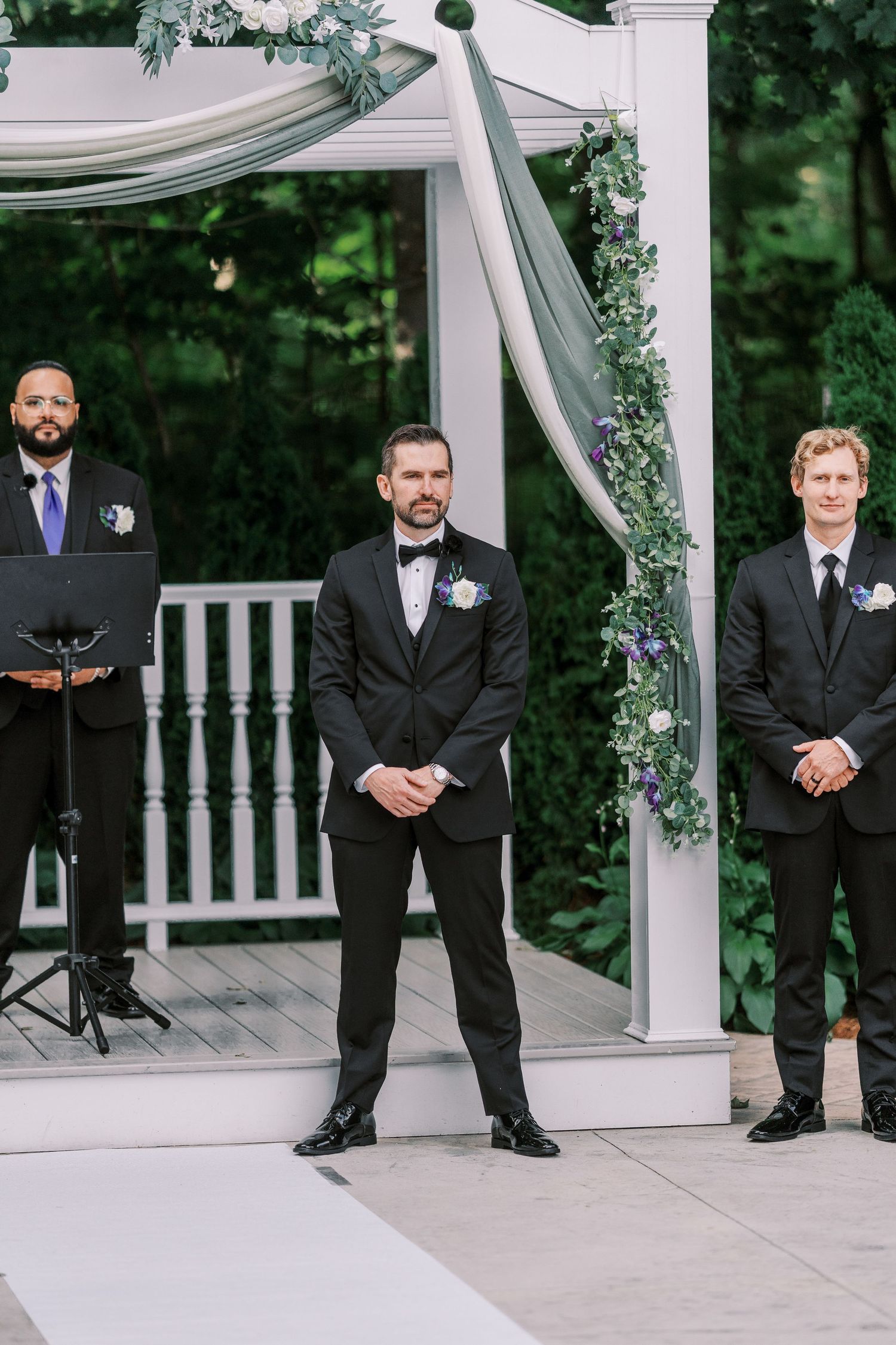 Groomsmen in black tuxedos stand at decorated wedding arch during outdoor ceremony at elegant white gazebo.