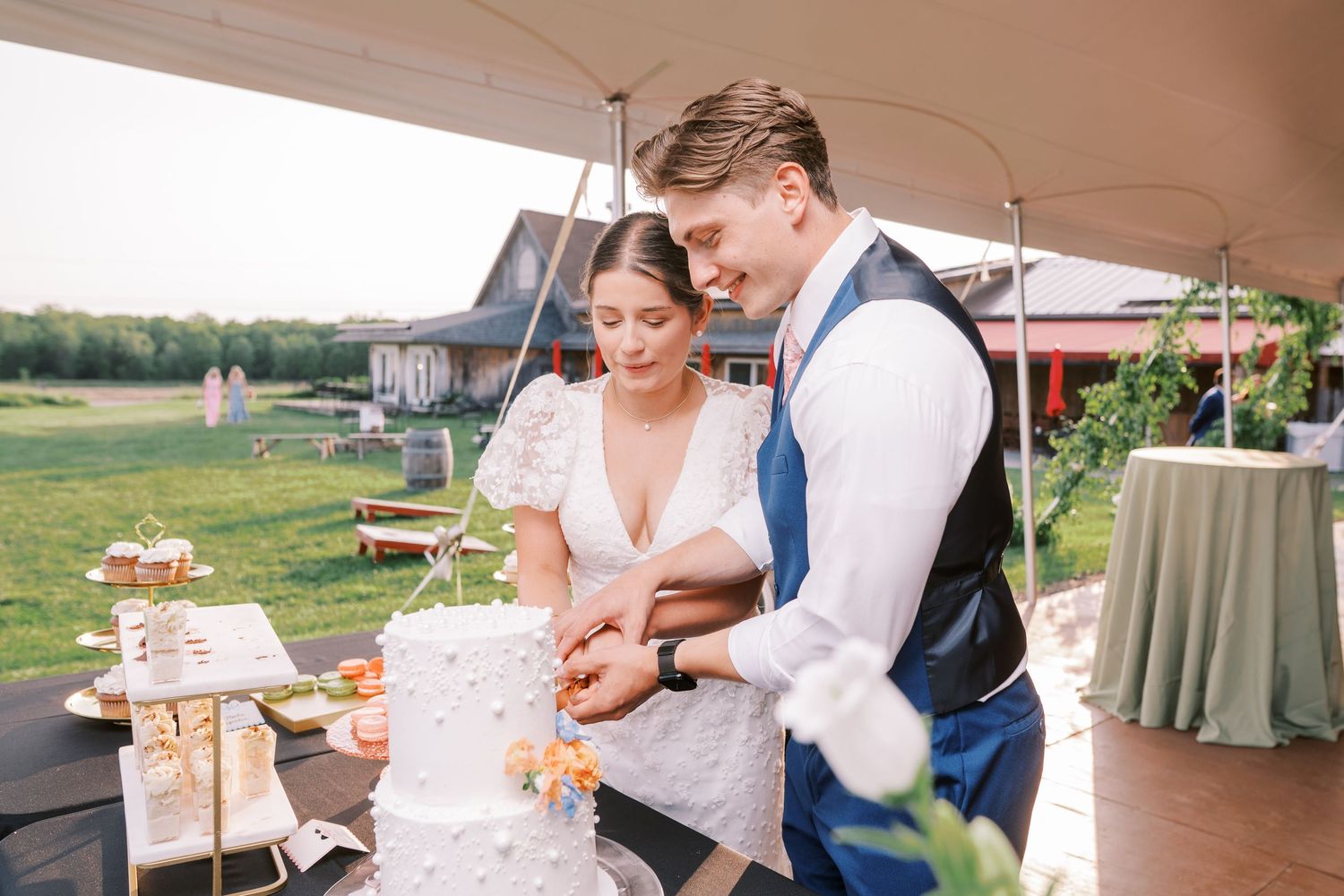 Couple cutting their elegant white wedding cake together under an outdoor tent during their countryside reception.