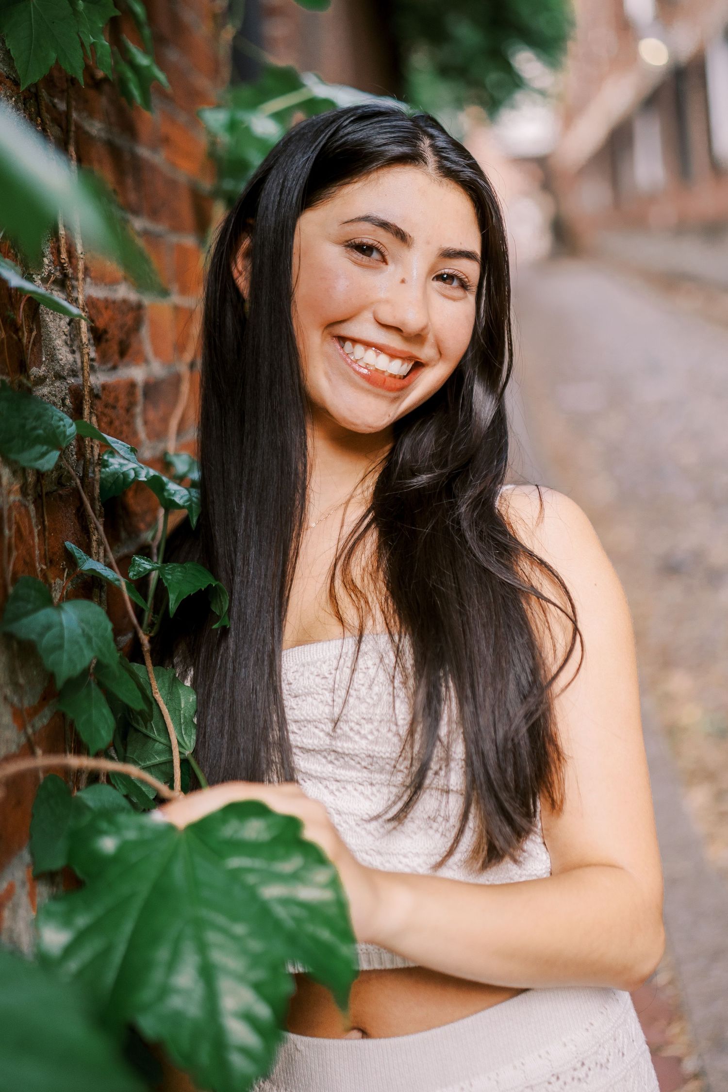 Portrait of someone with long dark hair wearing a white top smiling cheerfully against a brick wall with green ivy leaves.
