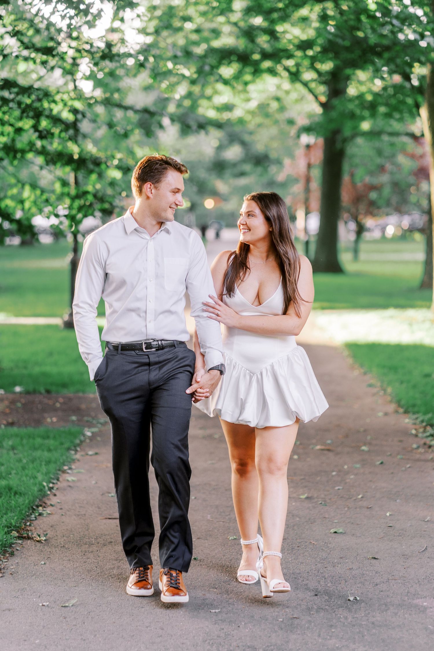 A couple in white and black outfits walks together on a tree-lined path in a park on a sunny day.