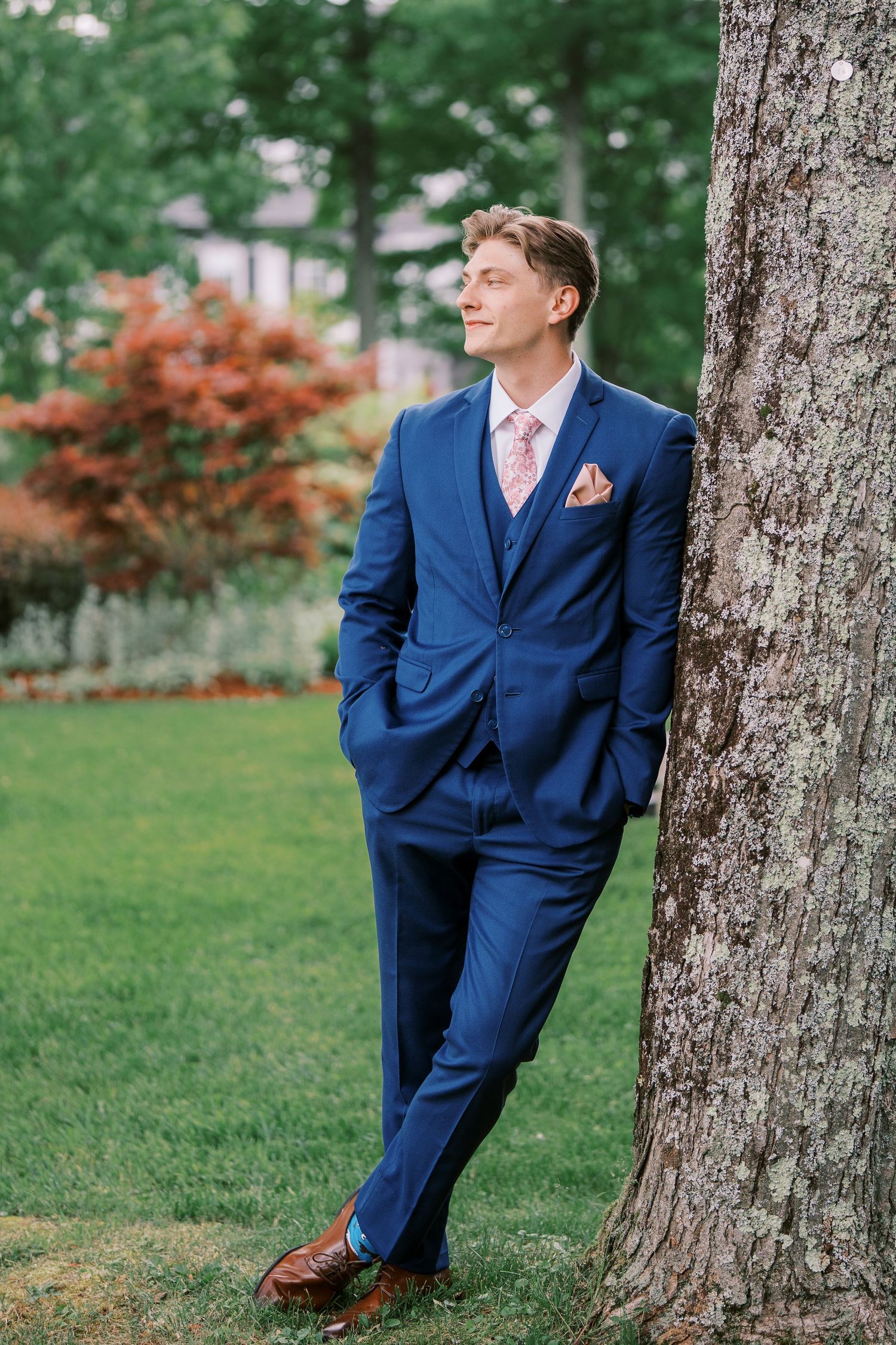 Person in navy blue suit and pink tie leans casually against large tree trunk in garden setting with red foliage.