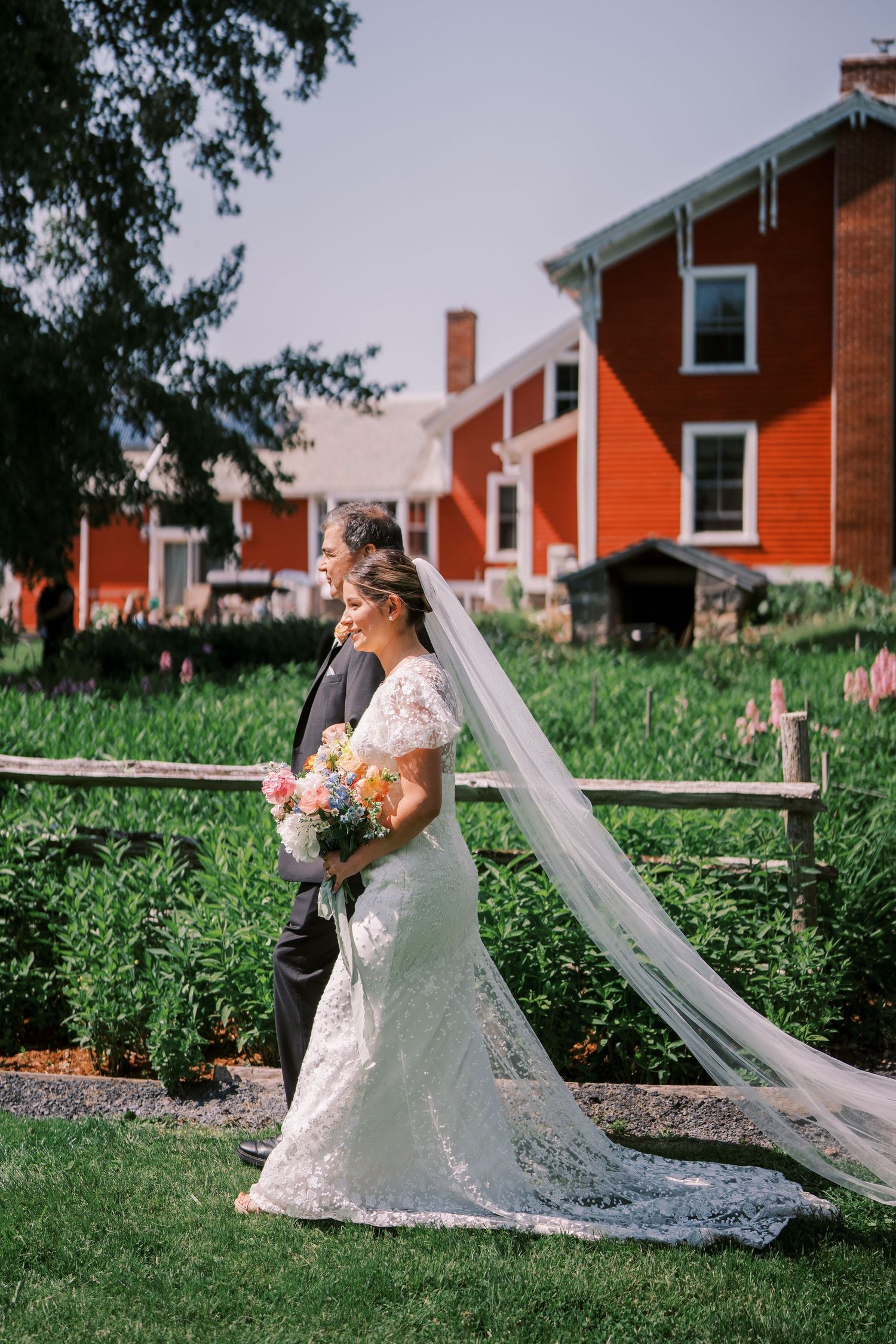 A couple in their wedding attire poses on a grassy lawn with red brick buildings in the background during a portrait session.