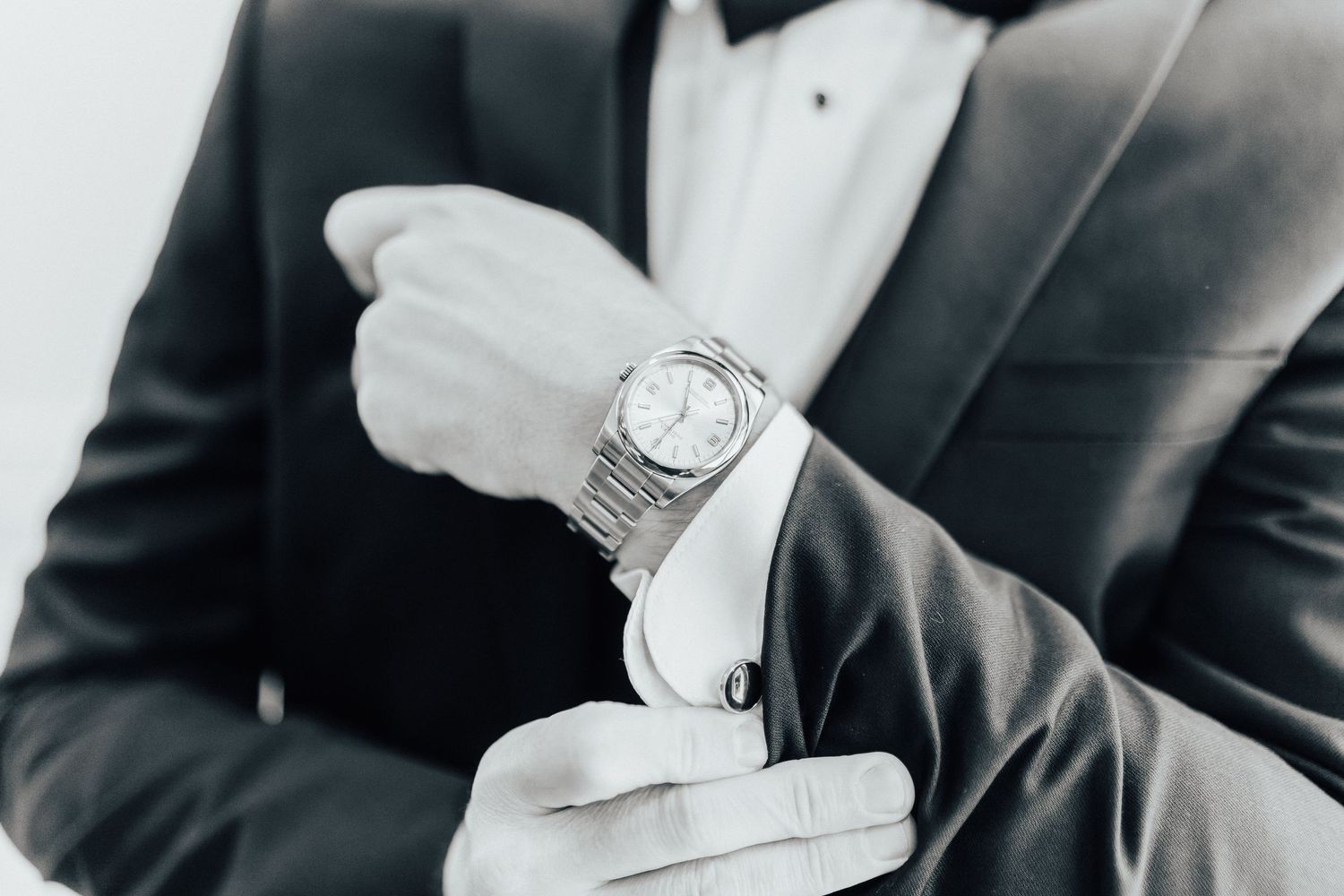 A person adjusts their wristwatch while wearing a formal black suit against a light background.