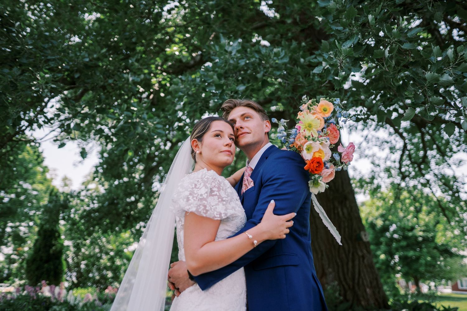 A romantic outdoor summer wedding scene with a couple embracing under lush green trees while holding bouquets.