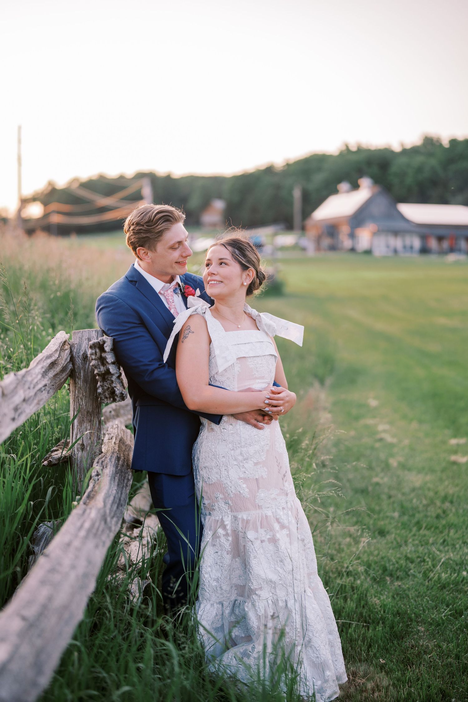 A couple in wedding attire share a romantic moment by a rustic wooden fence at sunset with a barn in the background.