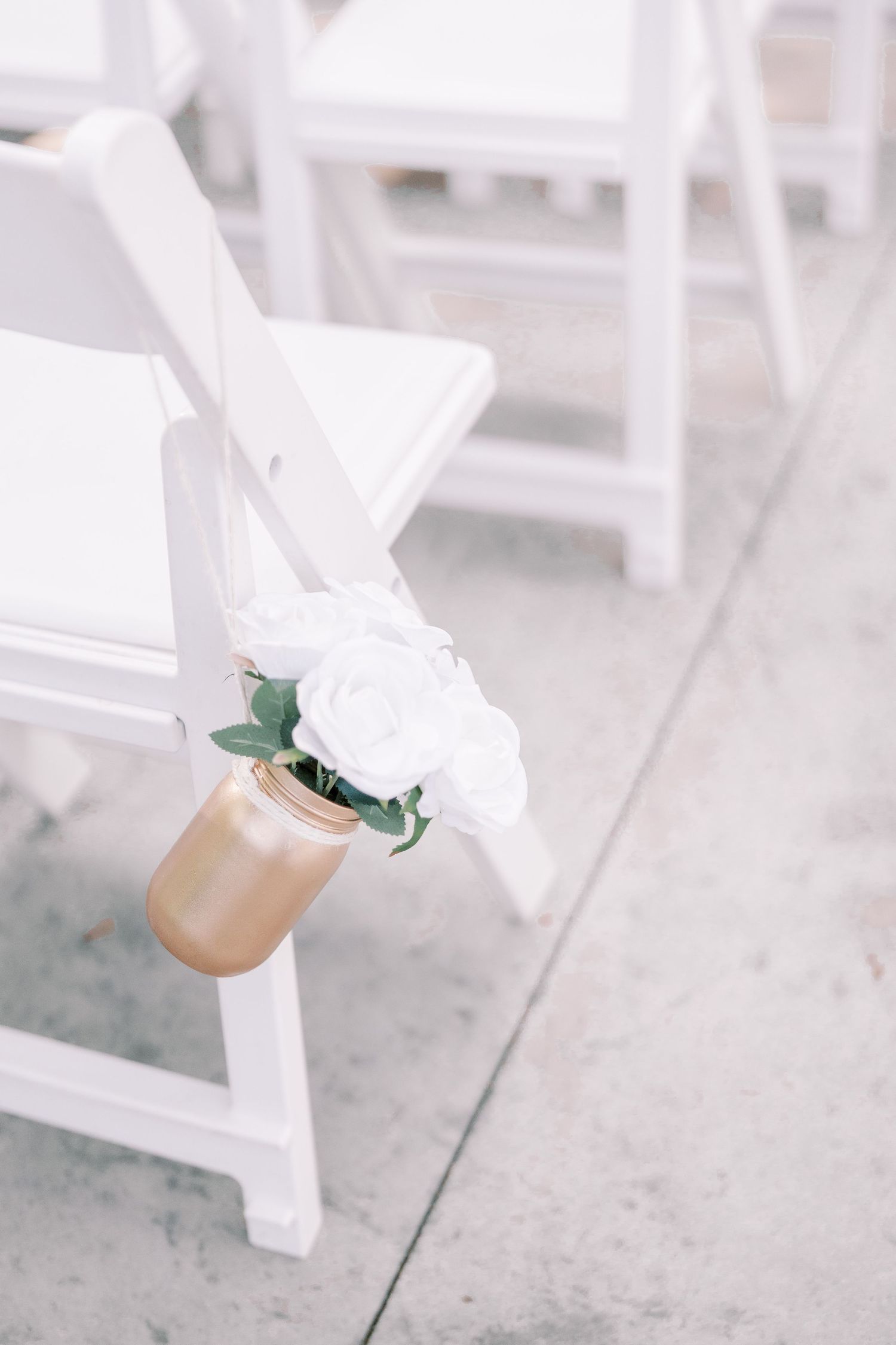 A white rose bouquet in a gold vase attached to a white ceremony chair at an elegant wedding venue.