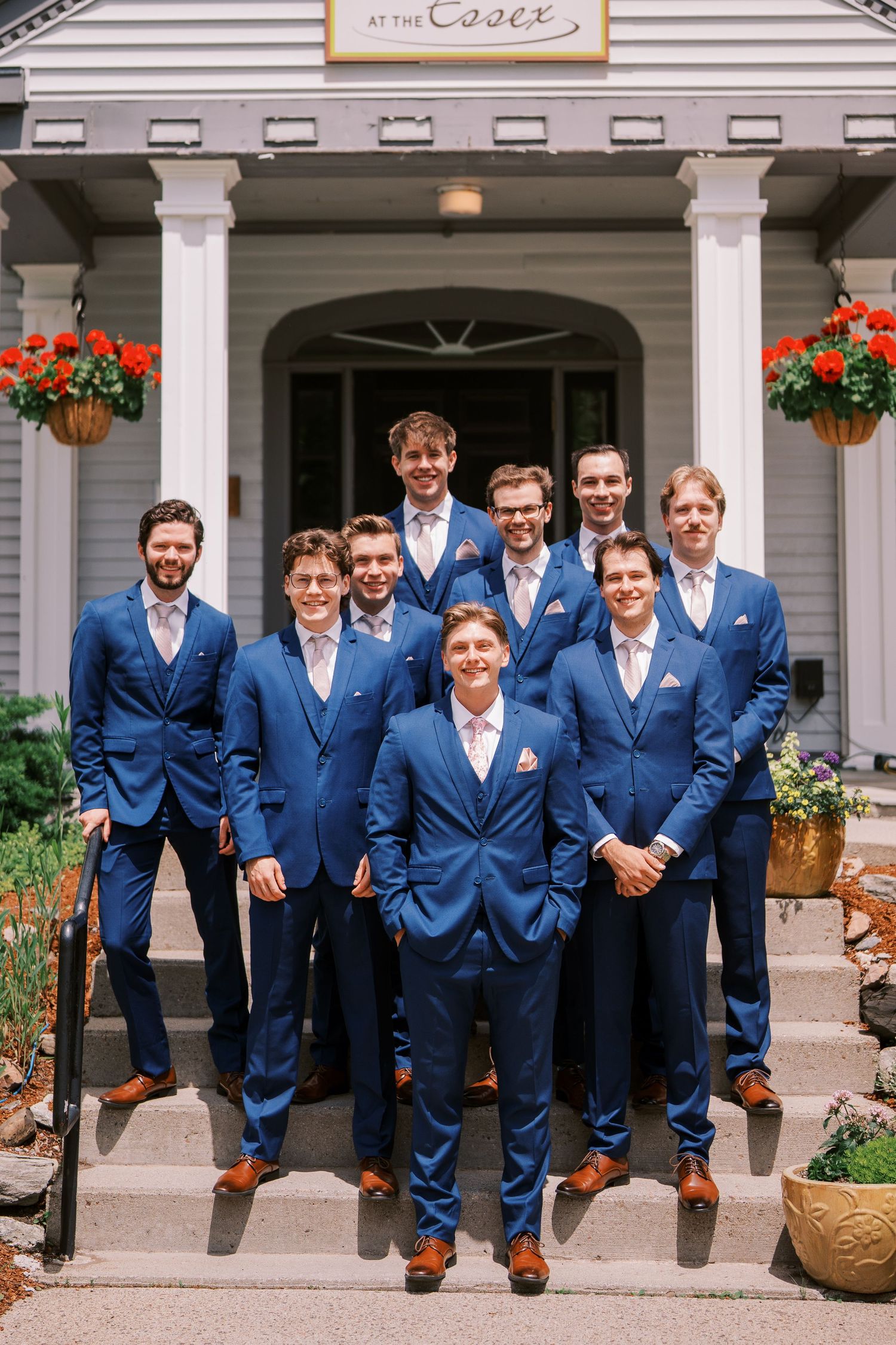A group of groomsmen in matching navy blue suits and brown shoes pose on the steps of a white-columned entrance.