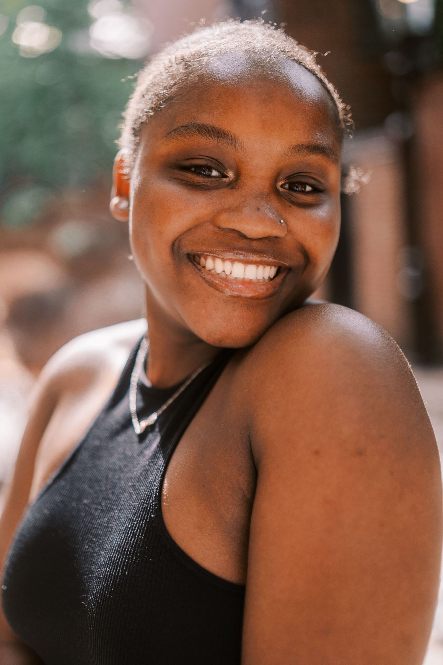 Portrait photo of someone wearing a black tank top smiling warmly at the camera with a blurred outdoor background.