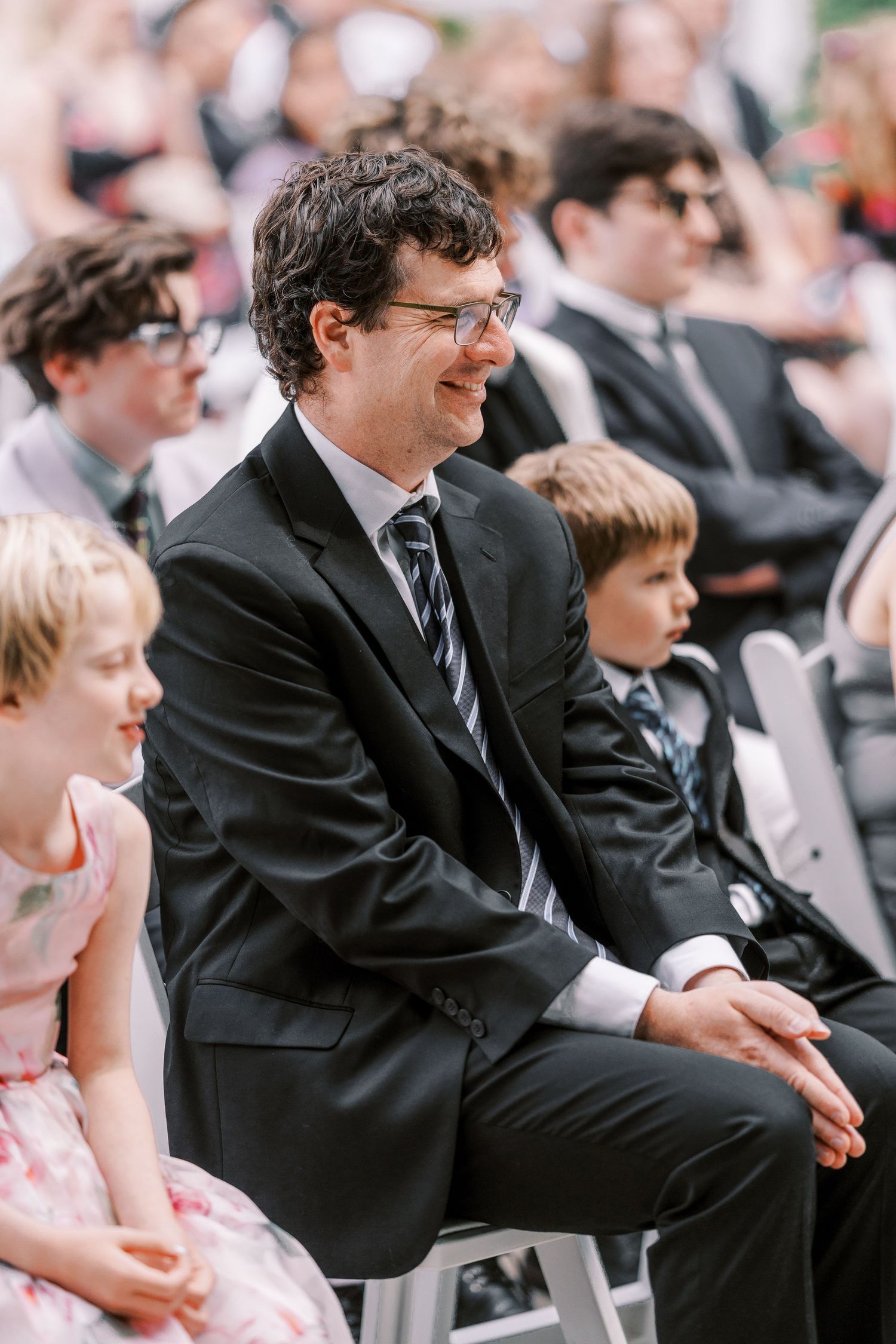 A guest in a black suit and striped tie sits and smiles at an outdoor event surrounded by other attendees.