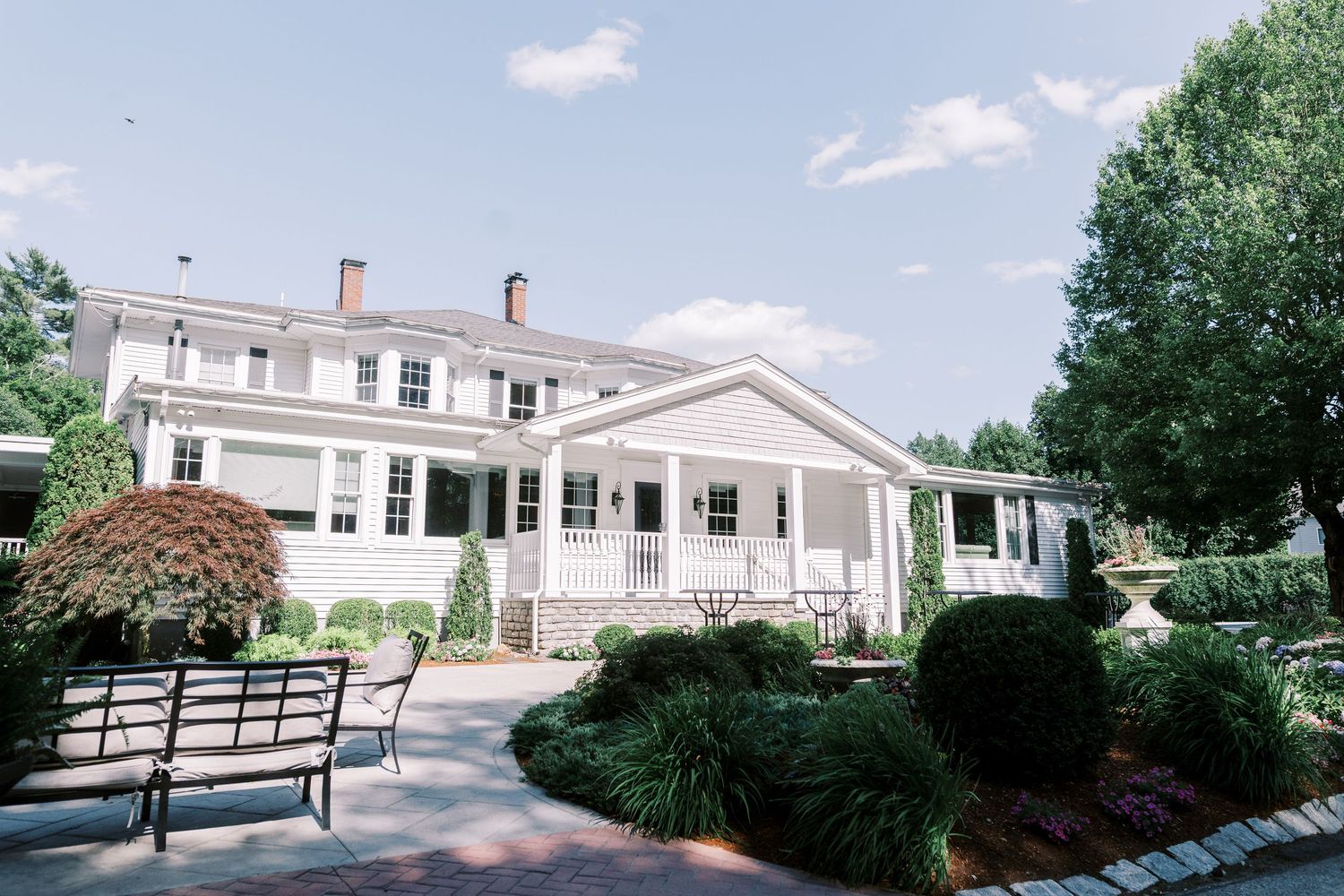 A set of white clapboard houses with green lawns are connected by a wooden walkway and benches under blue skies.