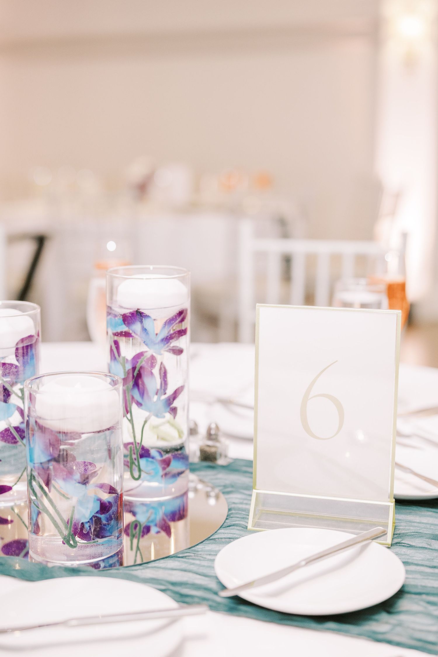 Wedding reception table setting with watercolor floral candle holders, teal tablecloth and elegant table number six display.
