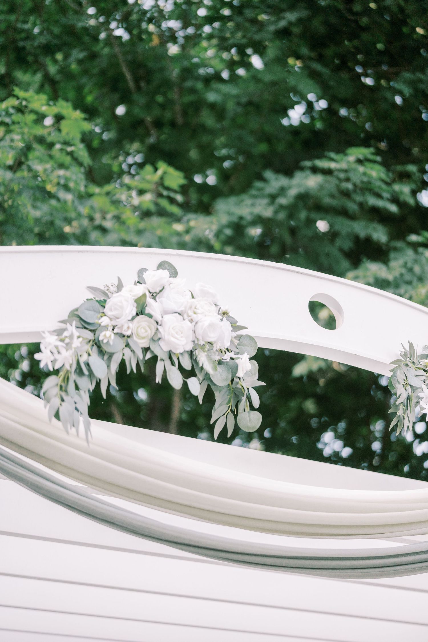 White floral arrangement with delicate roses and greenery decorates an elegant curved white bench at an outdoor wedding venue.