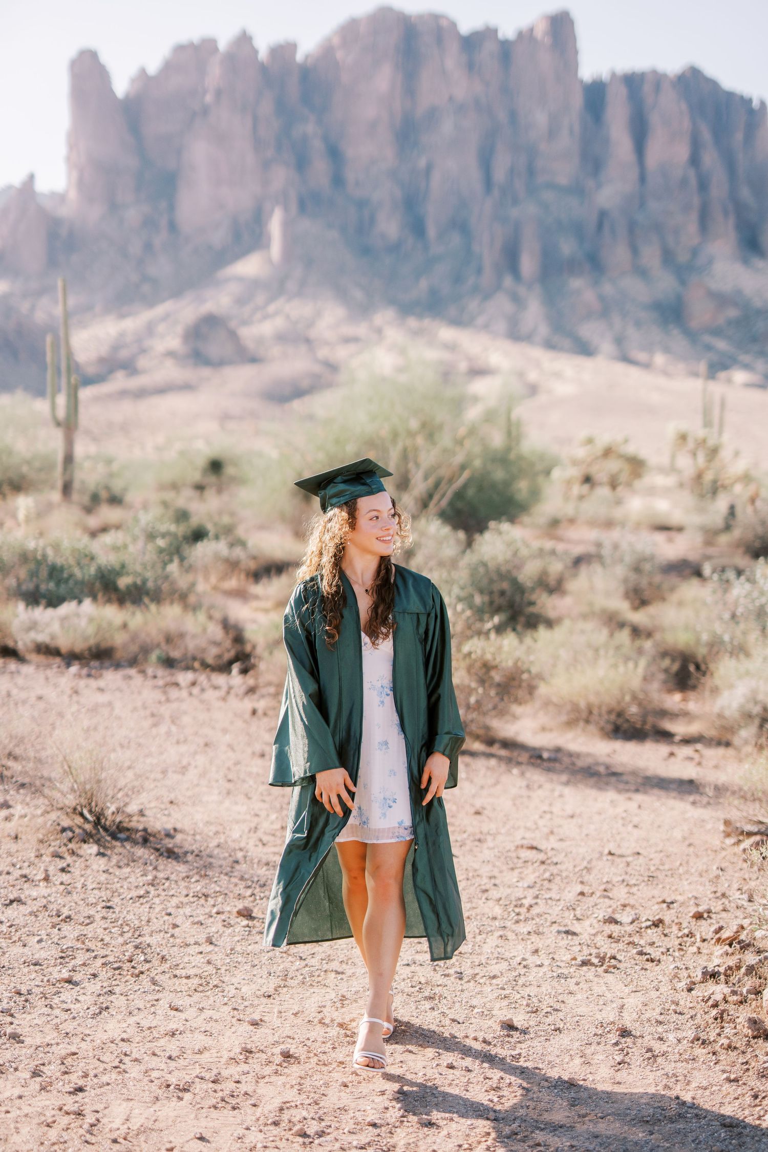 Graduate in green cap and gown poses in desert landscape with dramatic mountain cliffs and saguaro cacti in background.
