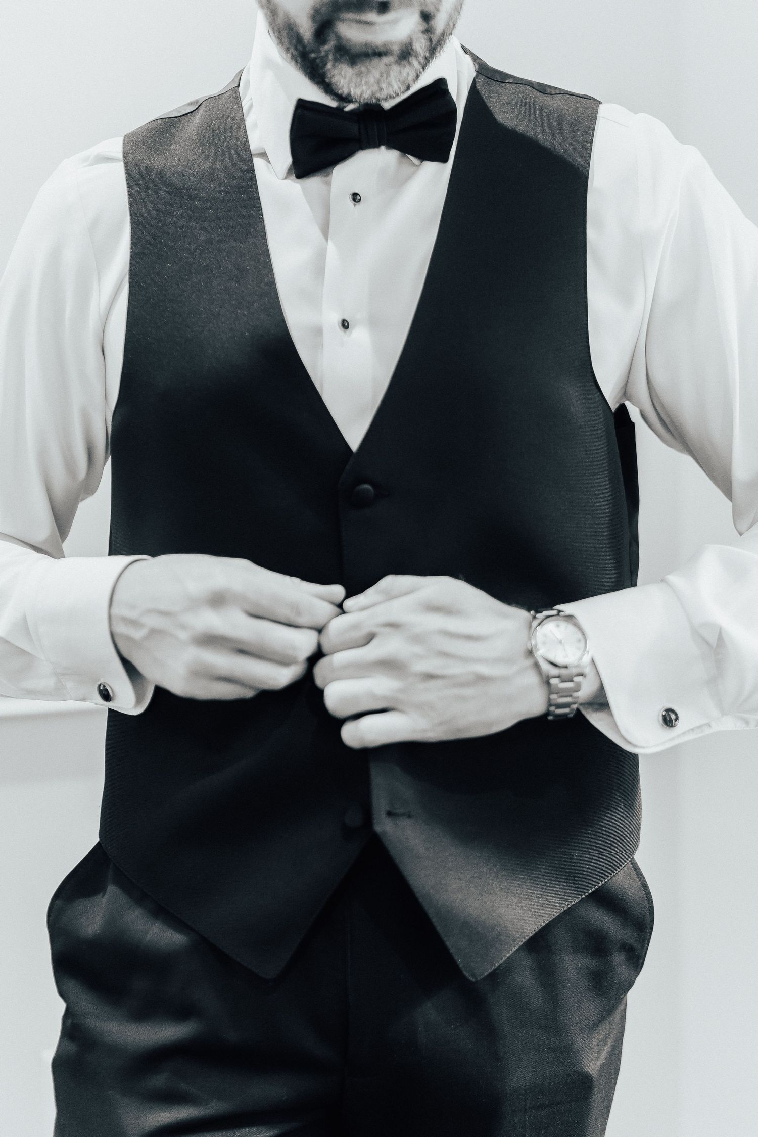 Close-up black and white photo of formal attire showing hands buttoning a dark vest over white dress shirt with bowtie.