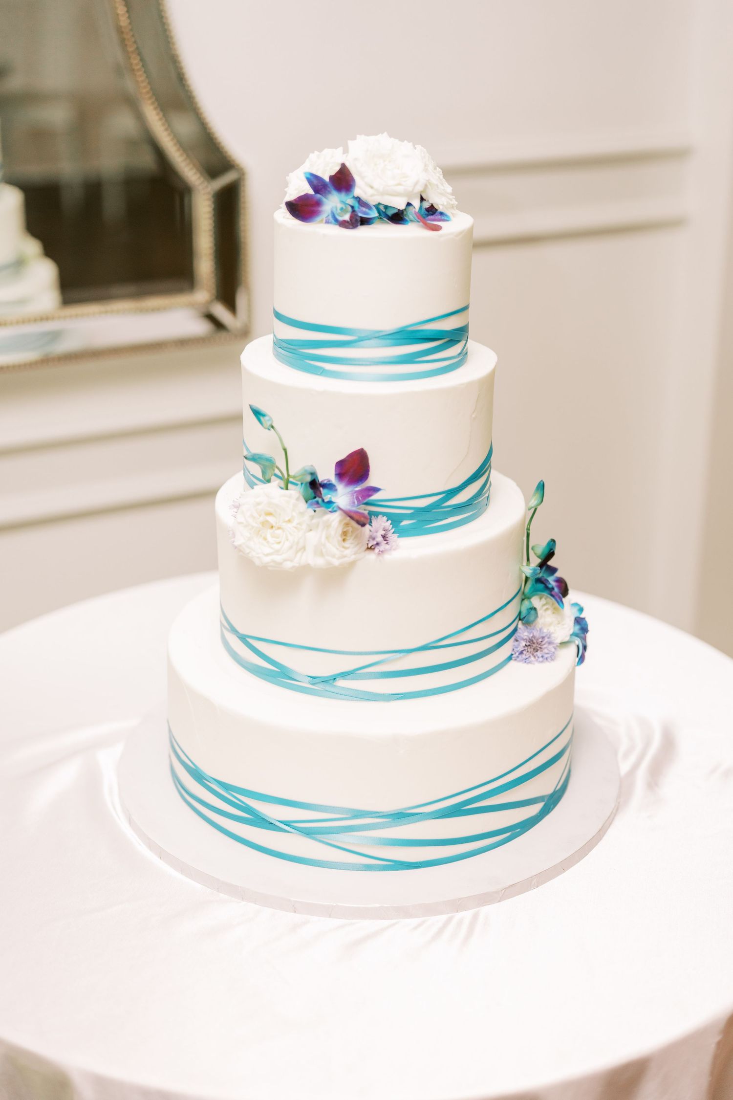 A four-tier white wedding cake decorated with turquoise ribbon-like stripes and purple and blue floral accents.