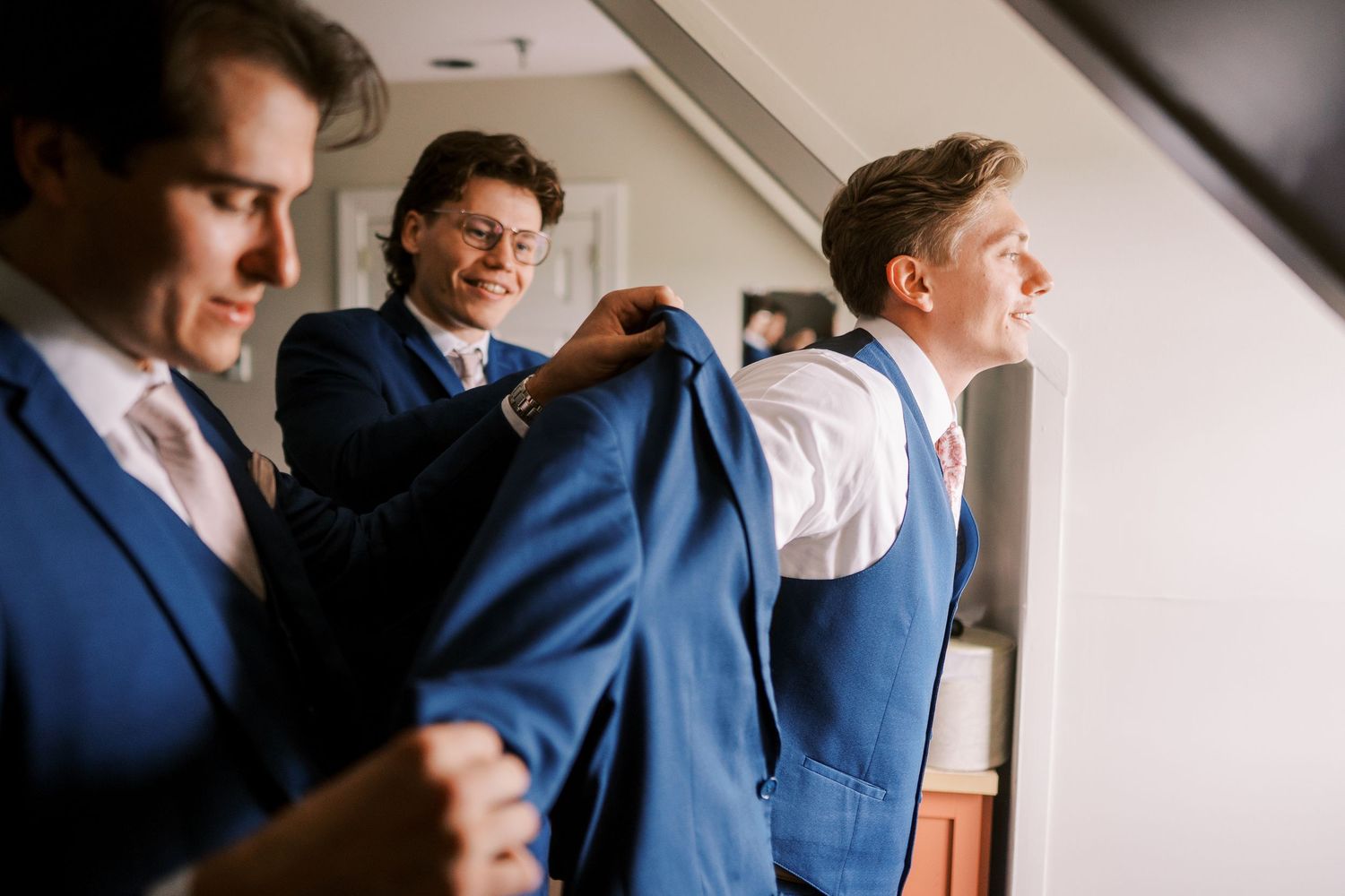 Groomsmen help each other get dressed in blue suits before a wedding celebration.