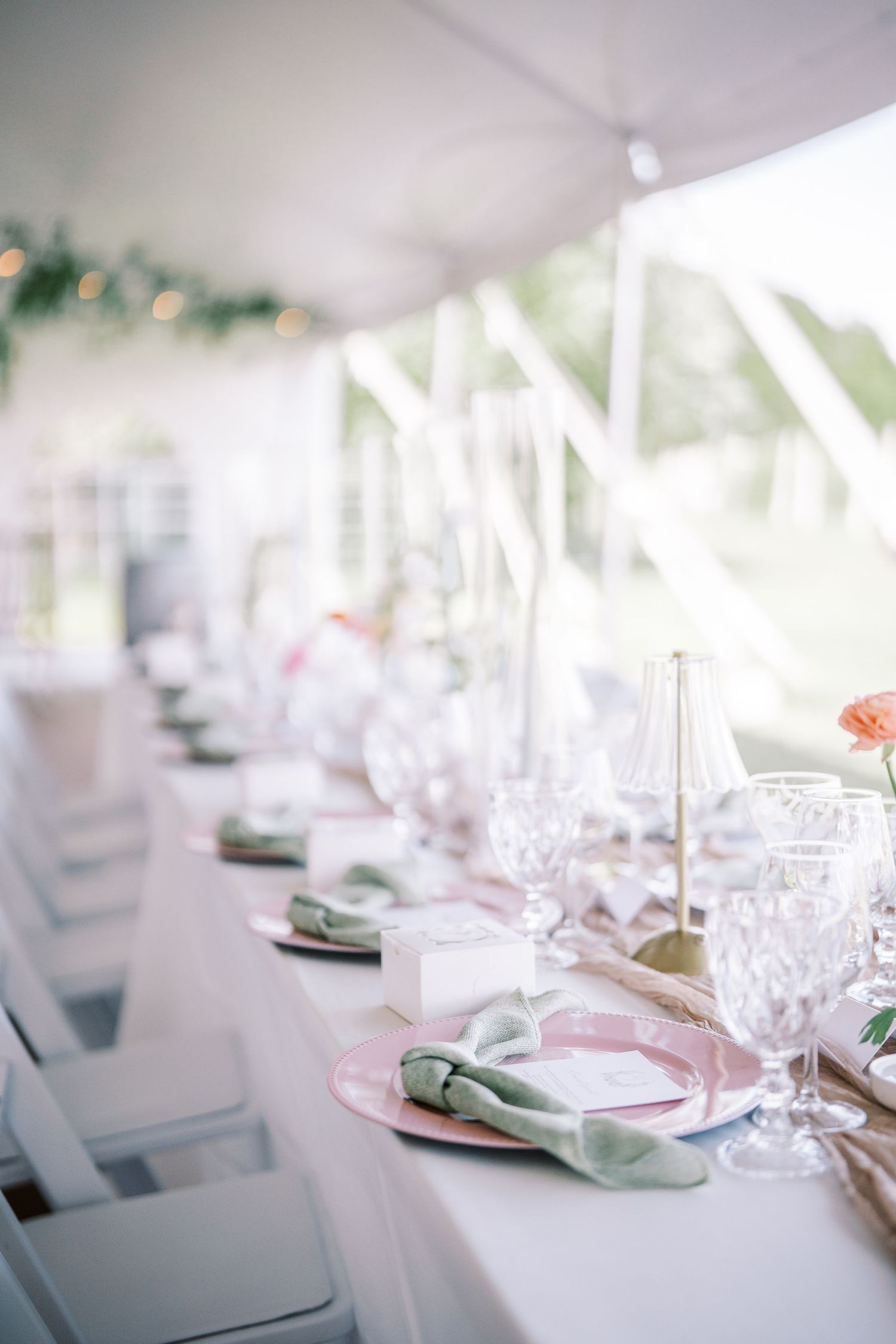 Elegant wedding reception table setting with crystal glassware, soft pink linens, and sage green napkins under a white tent.