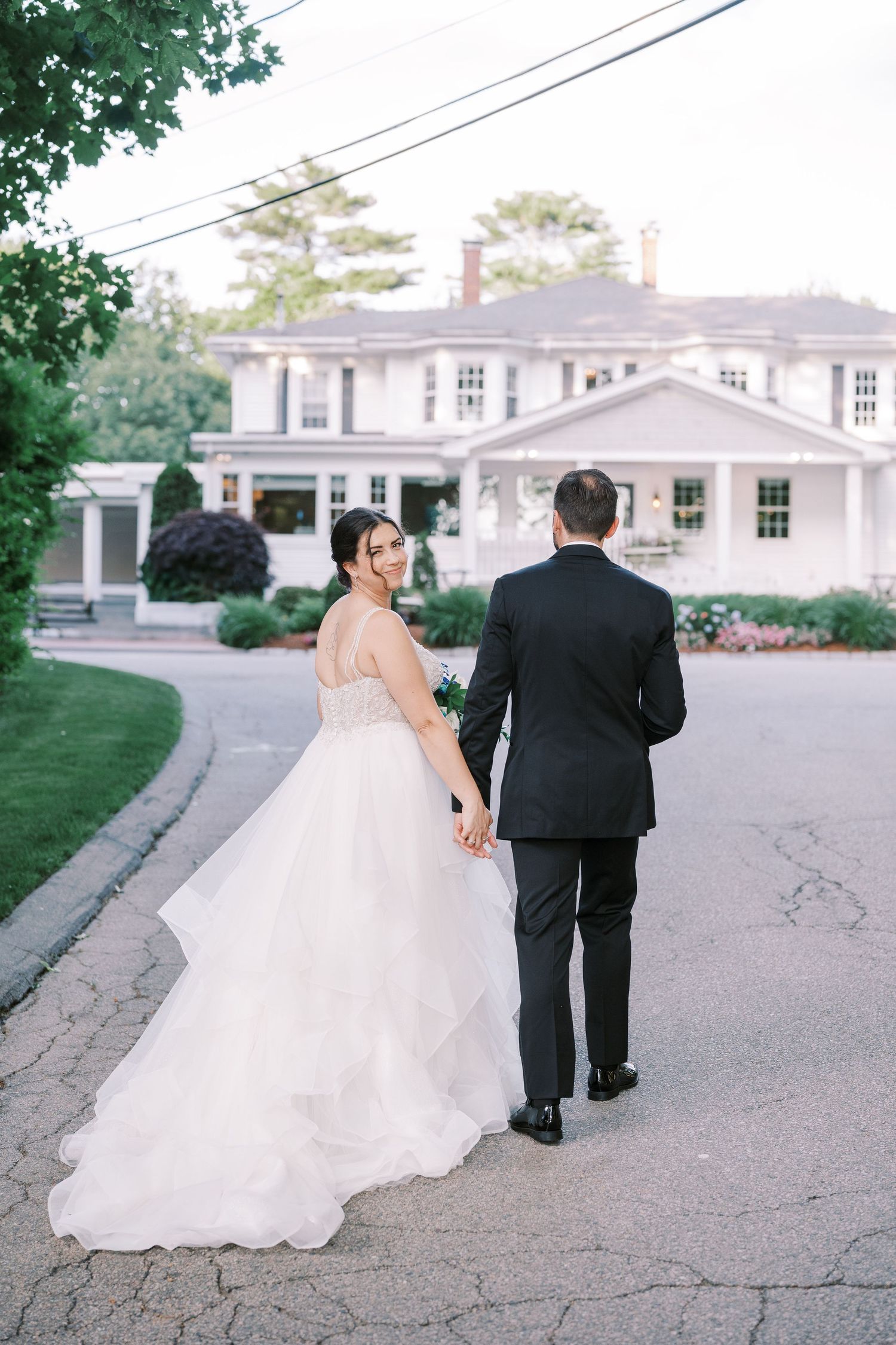 Wedding couple walks down a paved driveway surrounded by green landscaping at a white colonial-style venue.