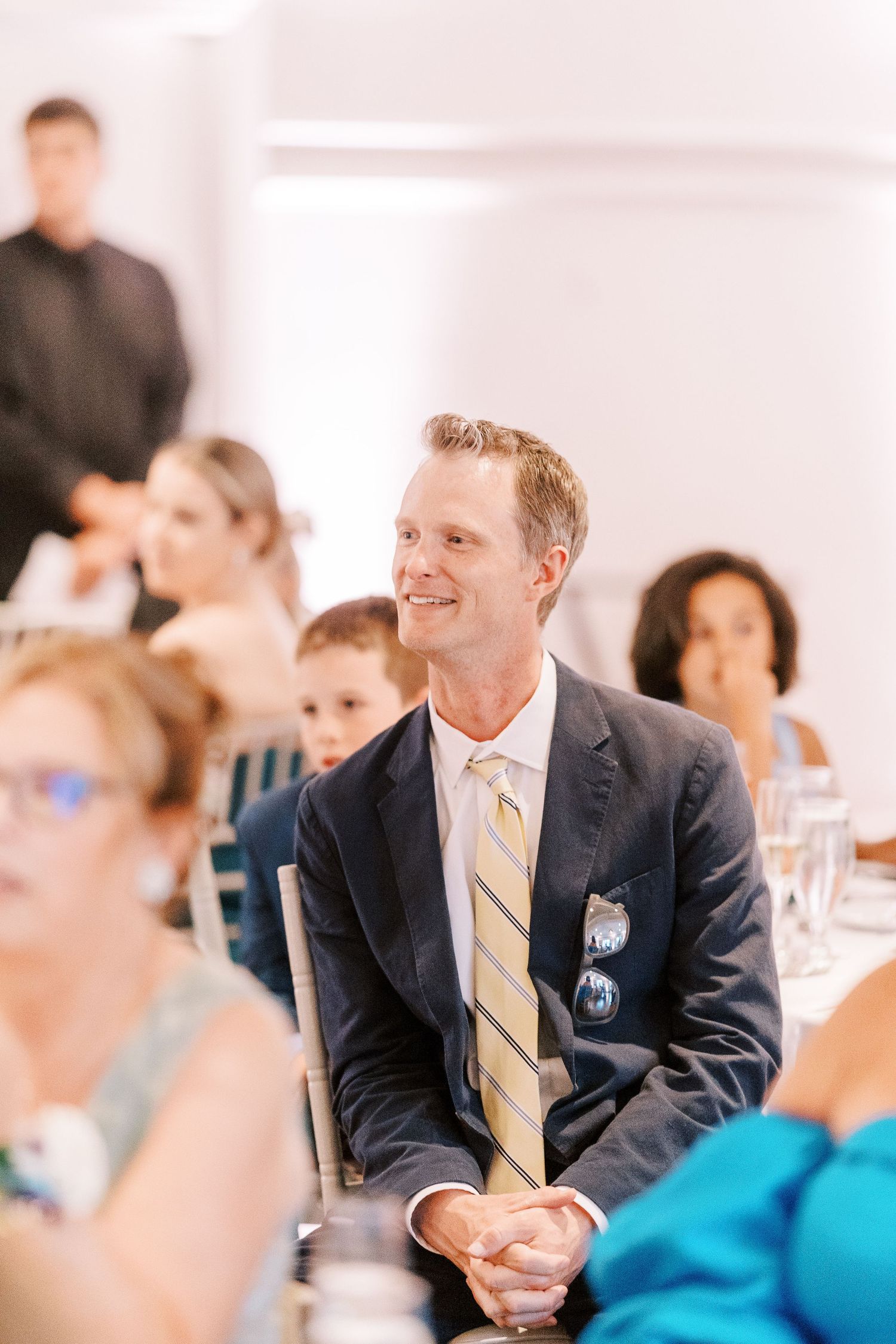 Wedding guest in navy suit and yellow striped tie smiles during reception dinner celebration.