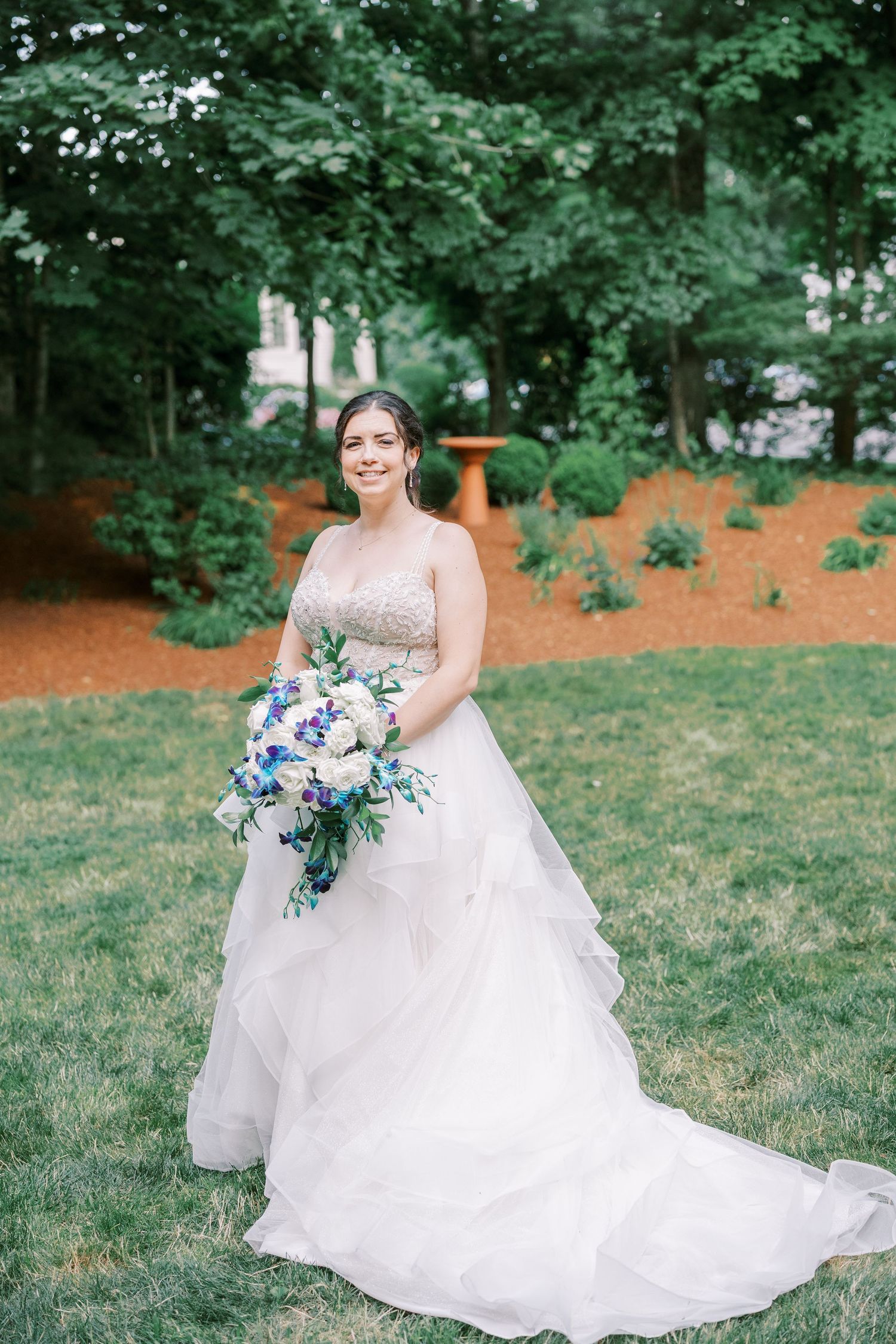 Bride in flowing white wedding dress sits gracefully on grass surrounded by lush green trees in outdoor garden setting.