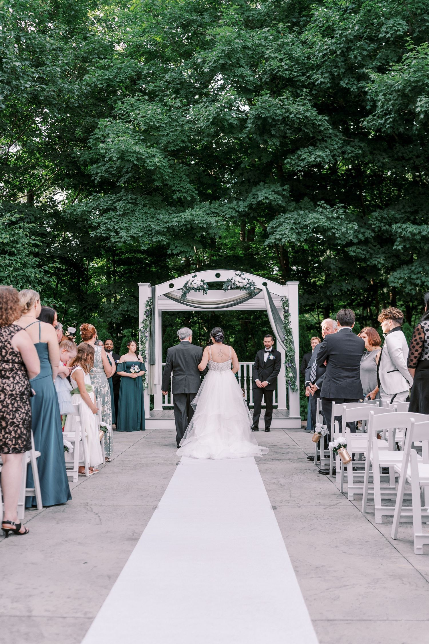 Wedding ceremony setup at an outdoor venue with wood arbor, white chairs, and lush green forest backdrop.