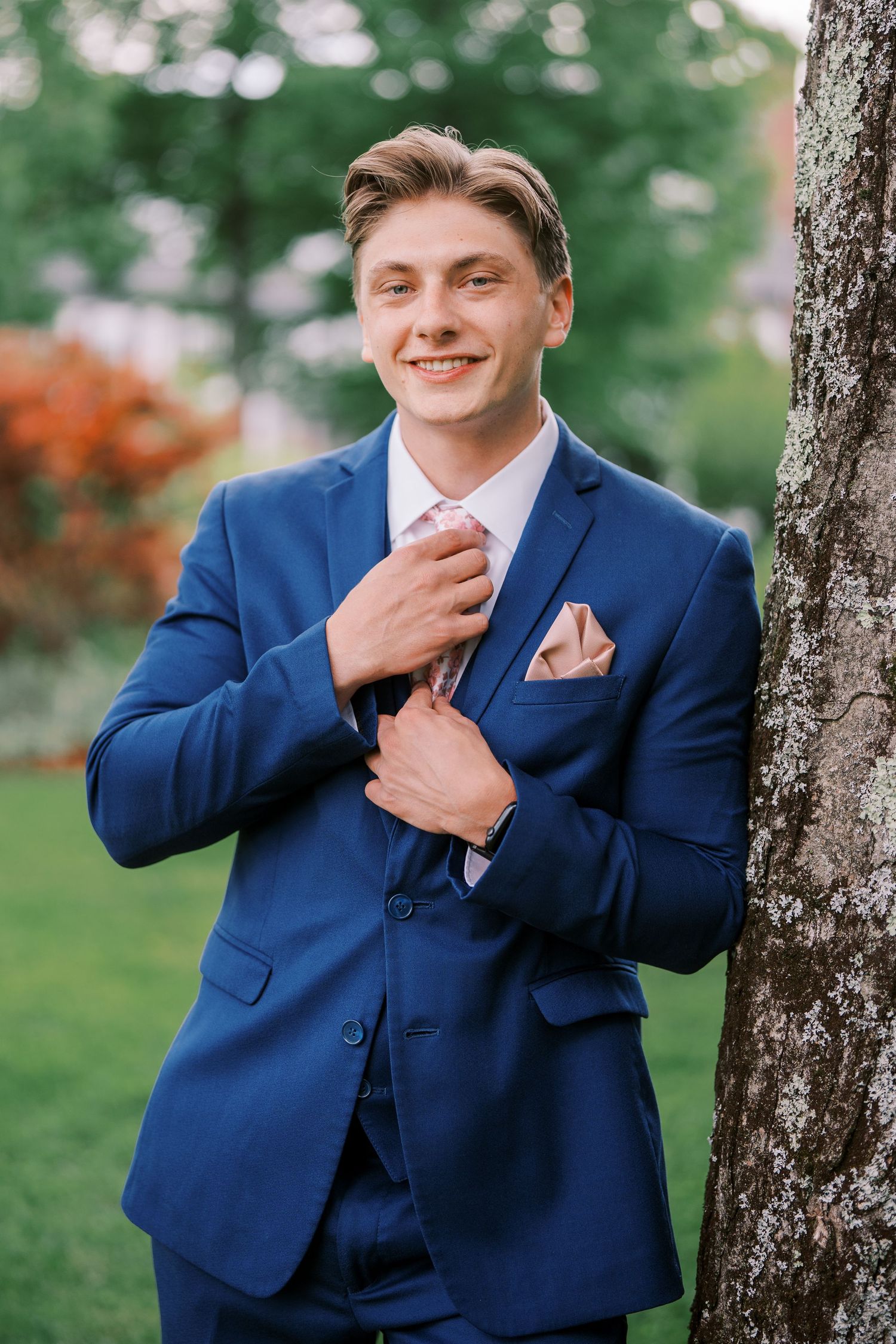 A person in a navy blue suit standing by a tree poses confidently in a garden setting.