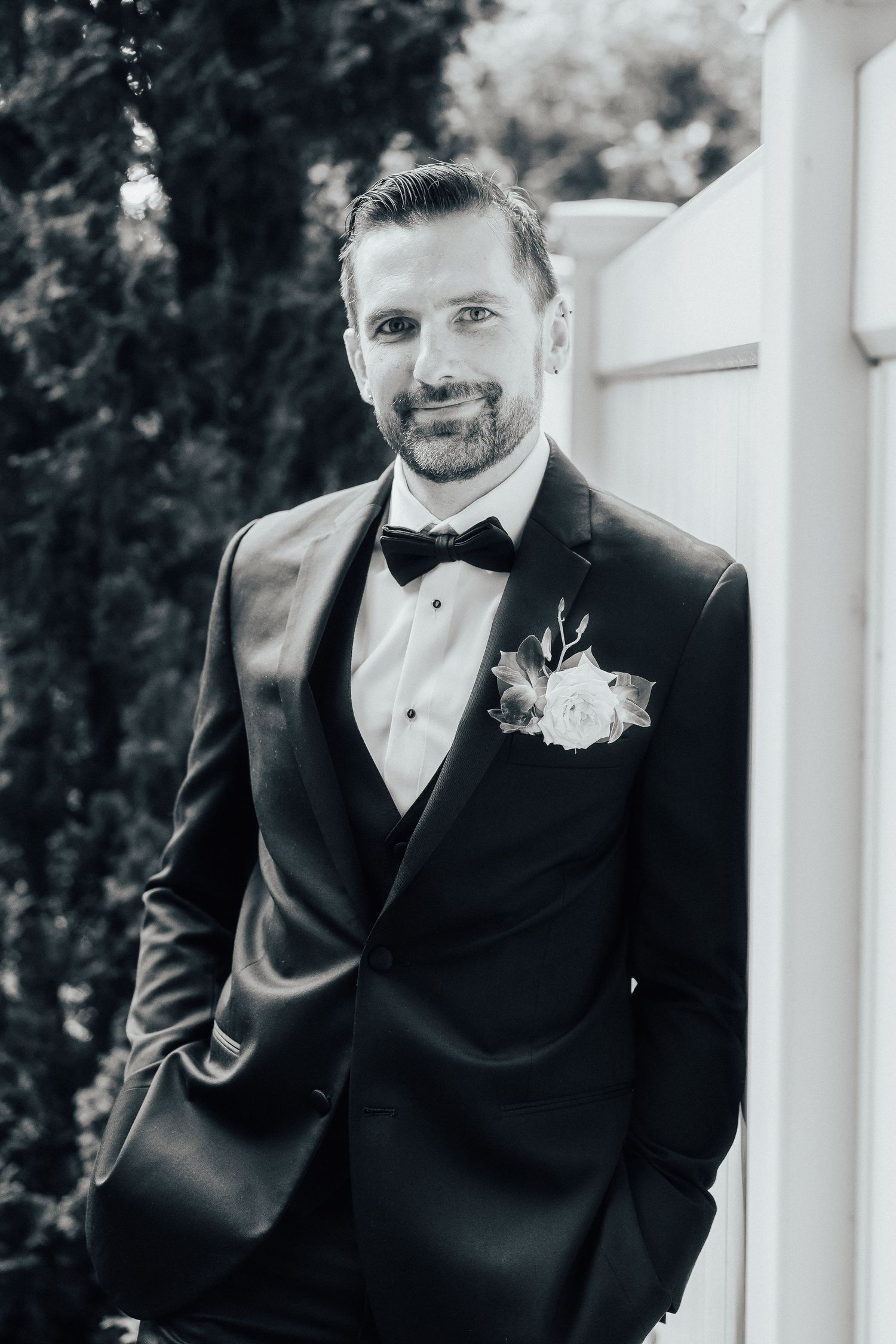 A groom in black tuxedo and bow tie smiles while standing against a white pillar at an outdoor wedding venue.
