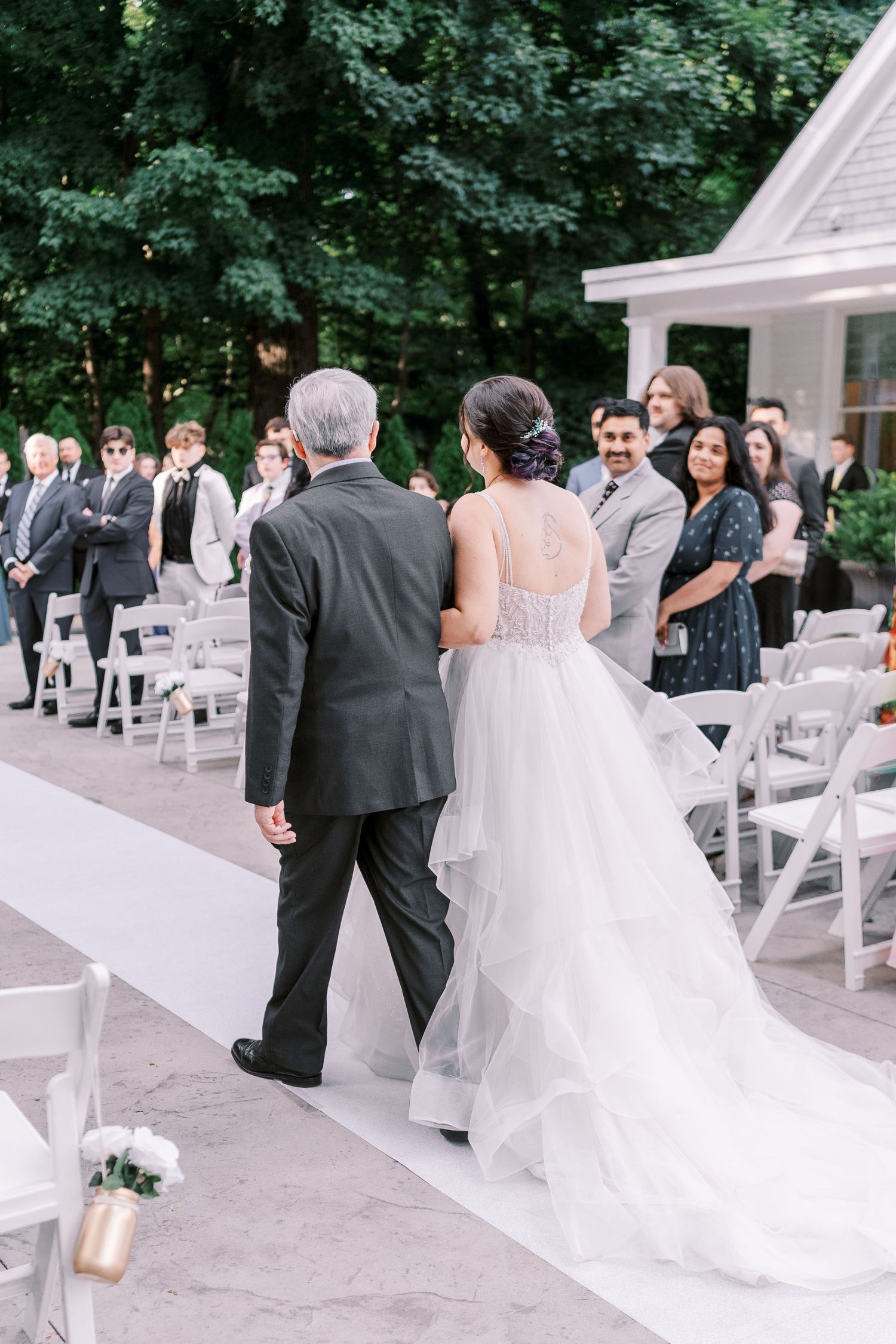 A bride in a white gown walks down an outdoor aisle lined with white chairs during an elegant garden wedding ceremony.