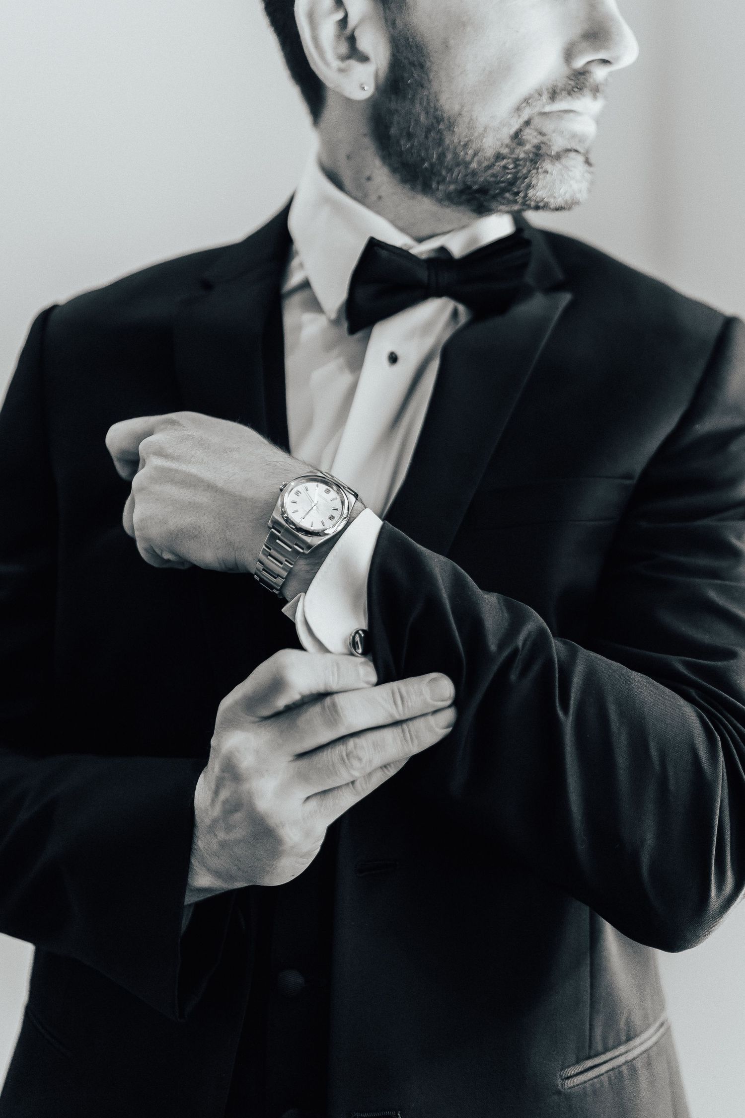 Close-up black and white shot showing someone adjusting a luxury watch while wearing a formal tuxedo with bow tie.