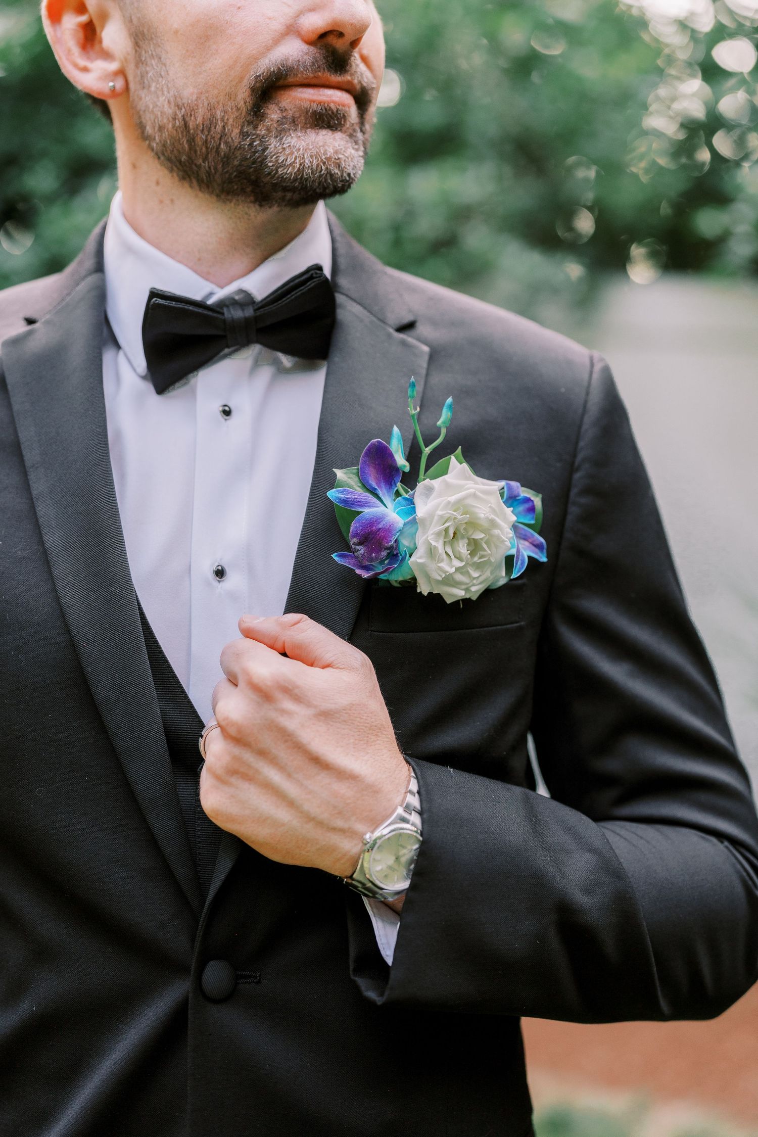 Close-up of groom in black tuxedo with blue orchid and white rose boutonniere and bow tie at wedding.